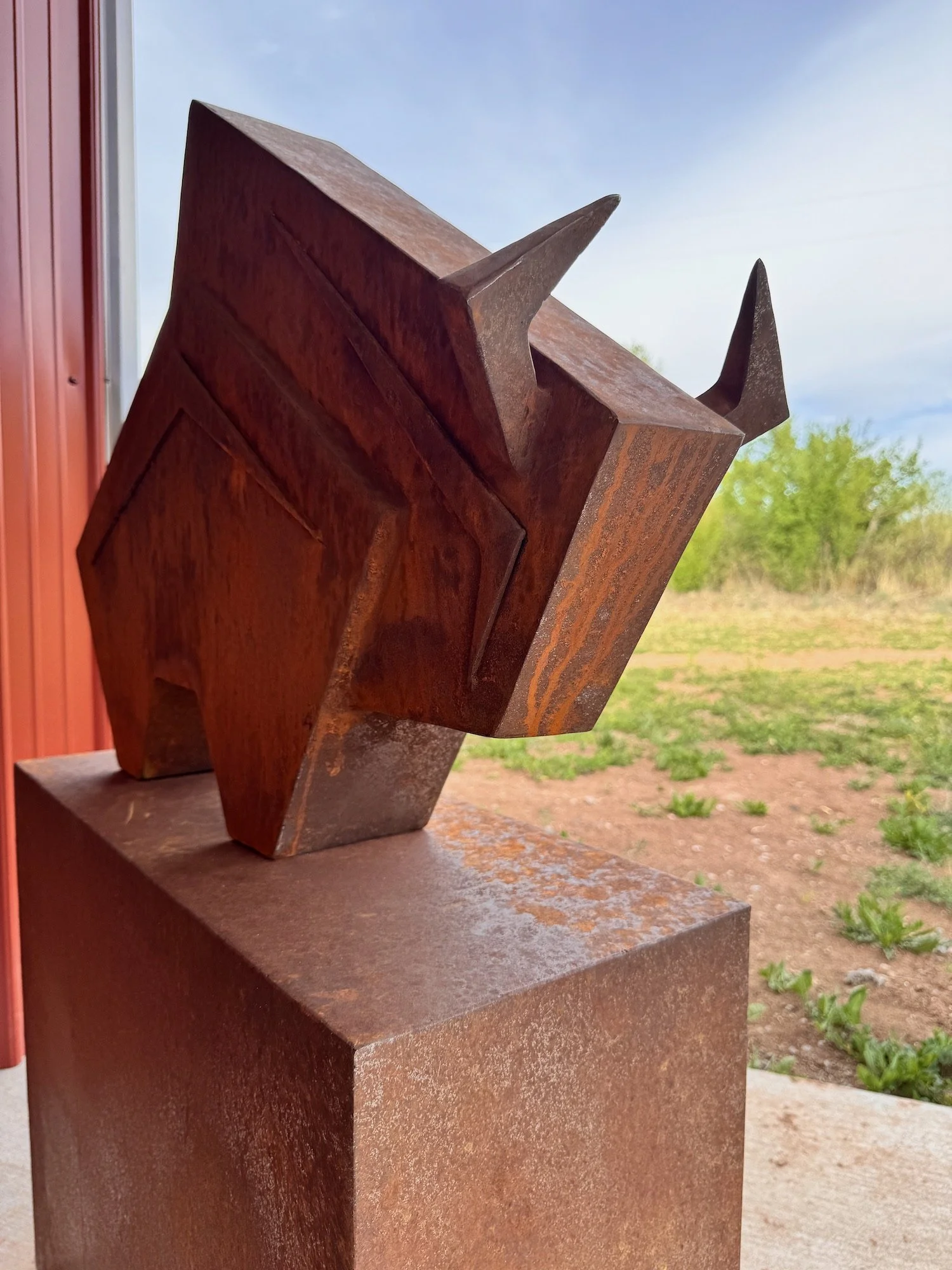 Close-up of a rusty metal sculpture with sharp, angular points, mounted on a rust-colored base, outdoors with a sandy ground, green bushes, and a blue sky in the background.