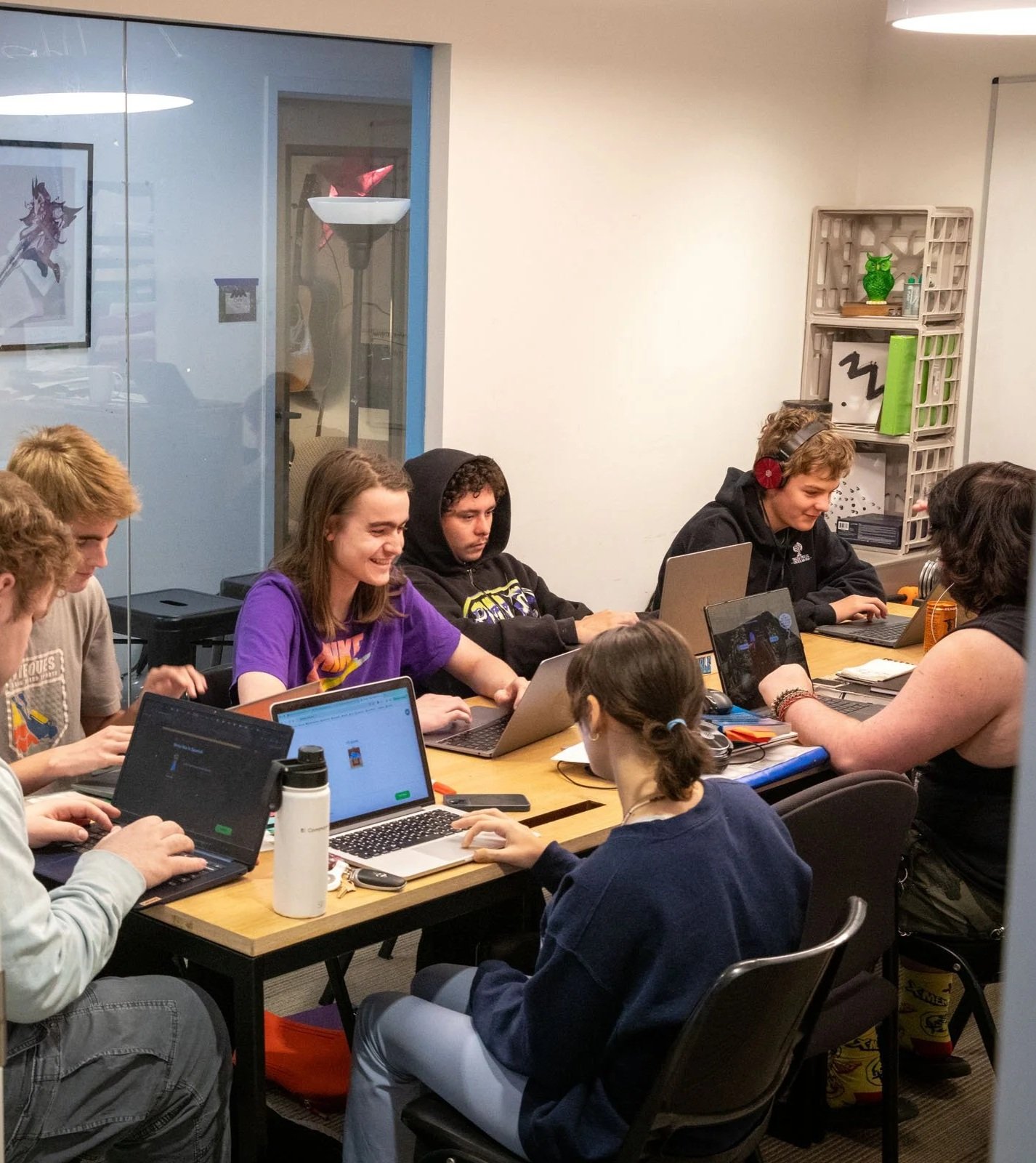 Group of young people working on laptops around a wooden table in a conference or classroom setting.