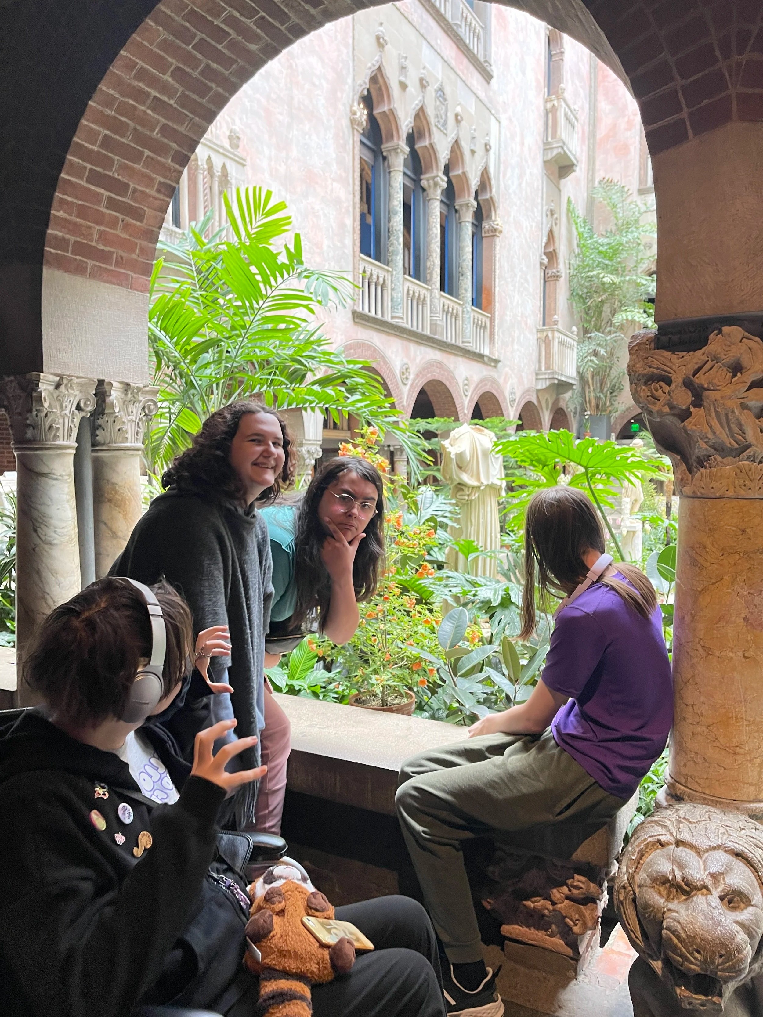 A group of people sitting and standing in an indoor garden area with lush green plants, ornate architectural details, and large windows in the background. Some individuals are engaged in conversation, and one person is looking at a phone.