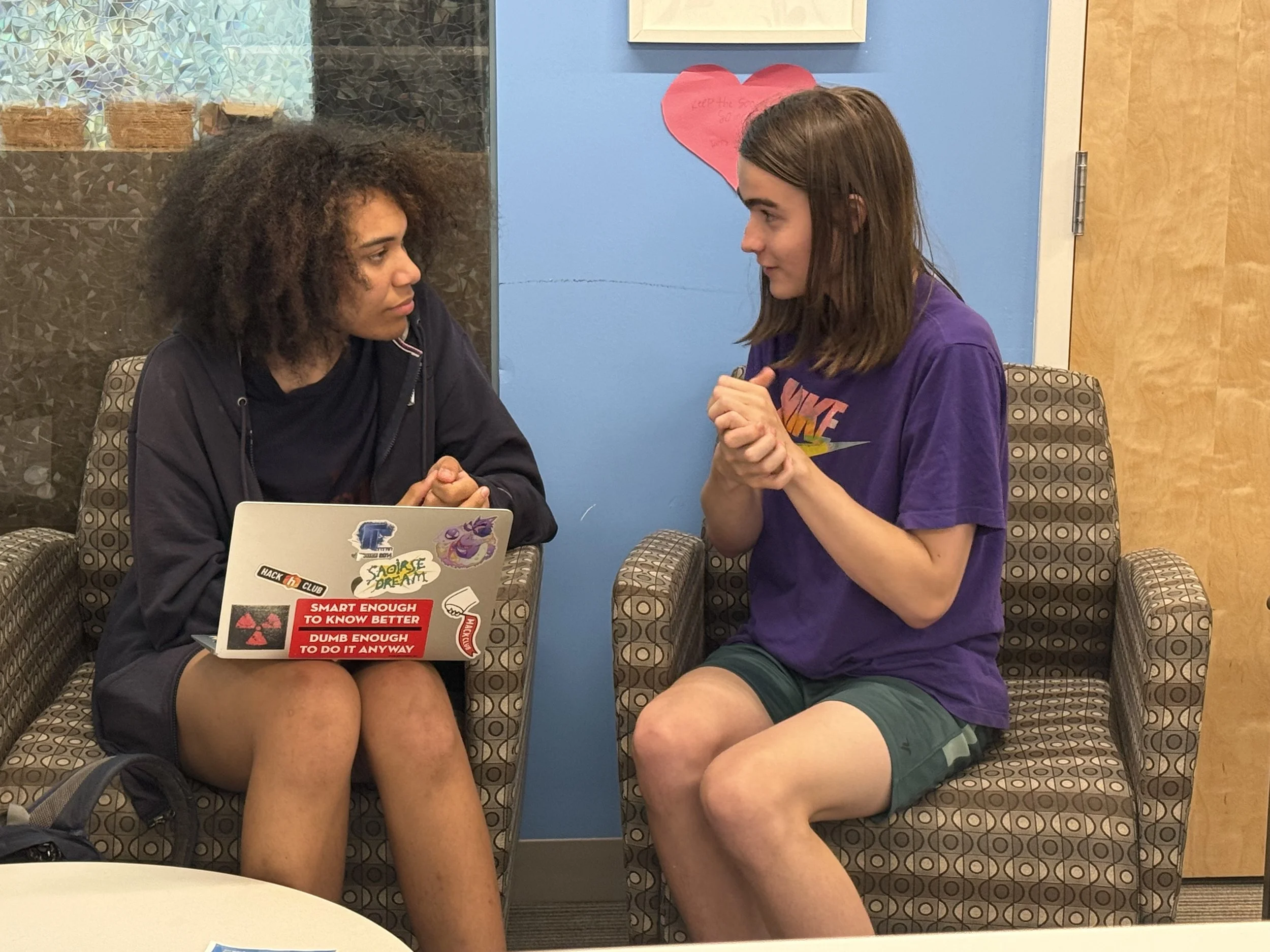 Two young people are sitting facing each other, engaged in conversation. The person on the left has curly hair and is wearing a dark hoodie, with stickers on the laptop in front of them that include phrases like 'Smart enough to know better' and 'Dumb enough to do it anyway.' The person on the right has straight brown hair, is wearing a purple t-shirt, and is gesturing with their hands. They are in a room with a blue wall, a wooden door, and a pink paper heart with writing on it in the background.