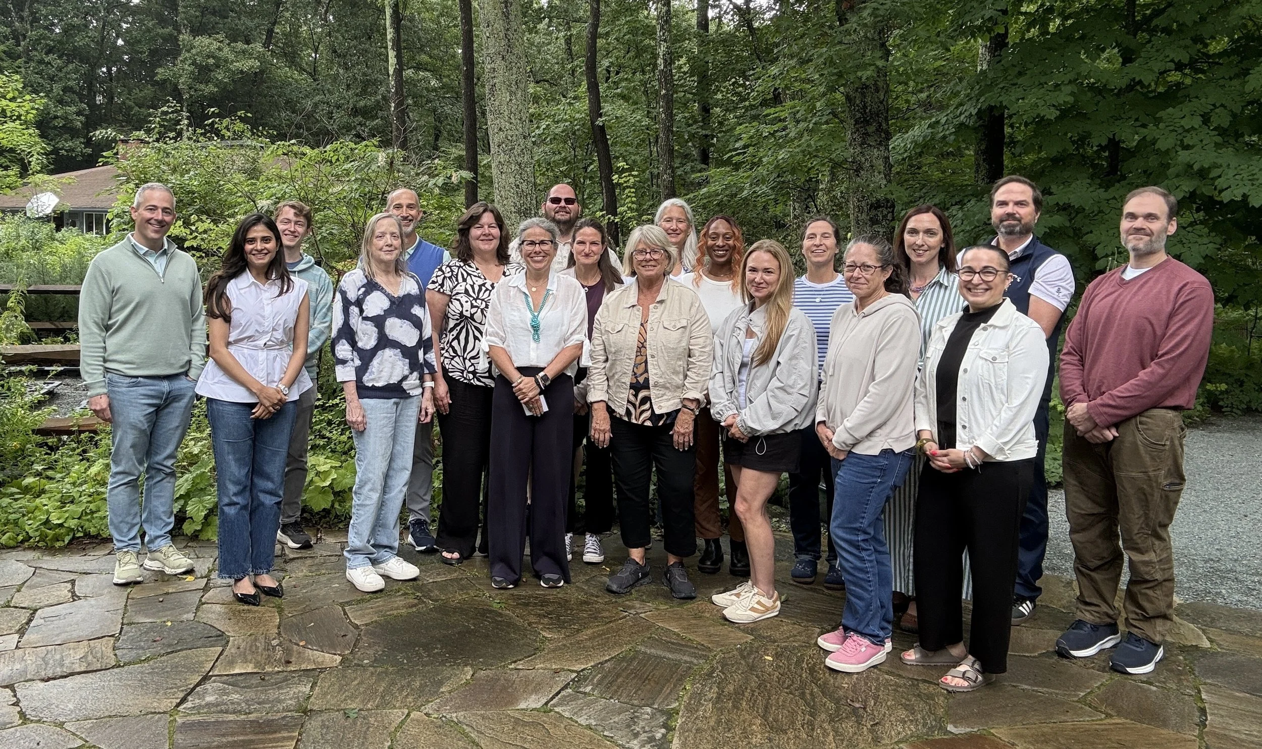 A group of 20 people standing outdoors on a stone patio in front of a wooded area, smiling and posing for a photo.