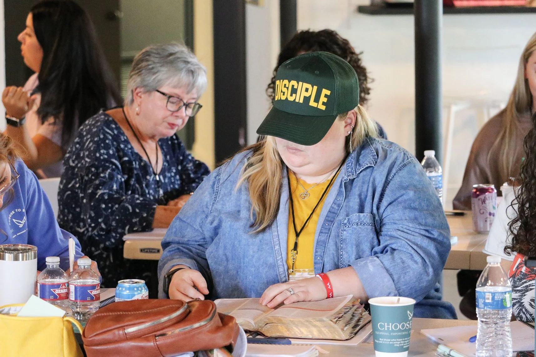 A woman with long blonde hair wearing a green cap with 'DISCIPLE' written on it, a denim jacket, and a yellow shirt, sitting at a table with various drinks and a book or notebook, in a crowded indoor setting.