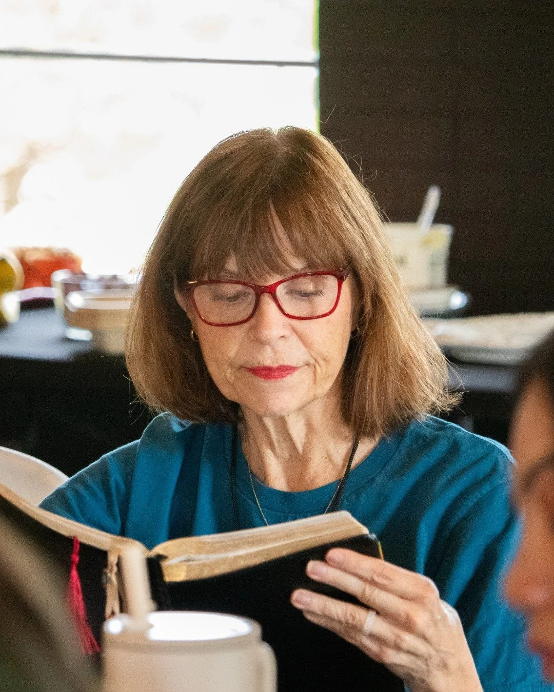 Older woman with red glasses reading a book at a table, with food and condiments in the background.