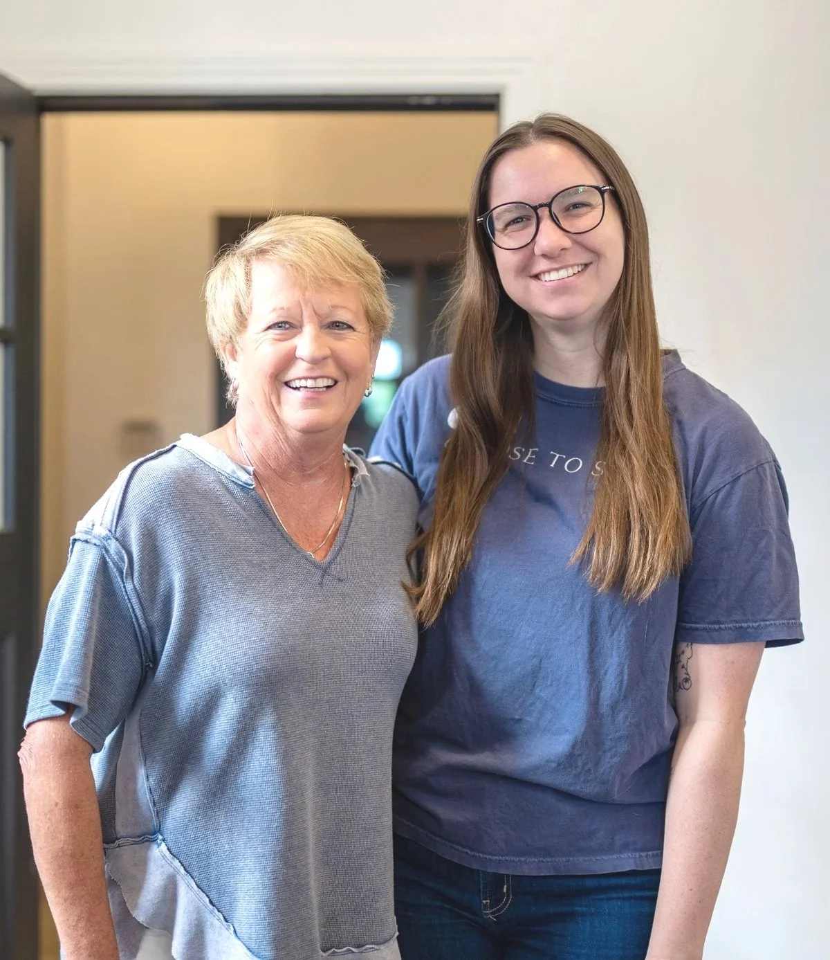 Two women smiling indoors, standing close. The older woman has short, light-colored hair and wears a grey, loose-knit top. The younger woman has long, brown hair, glasses, and wears a dark blue T-shirt.