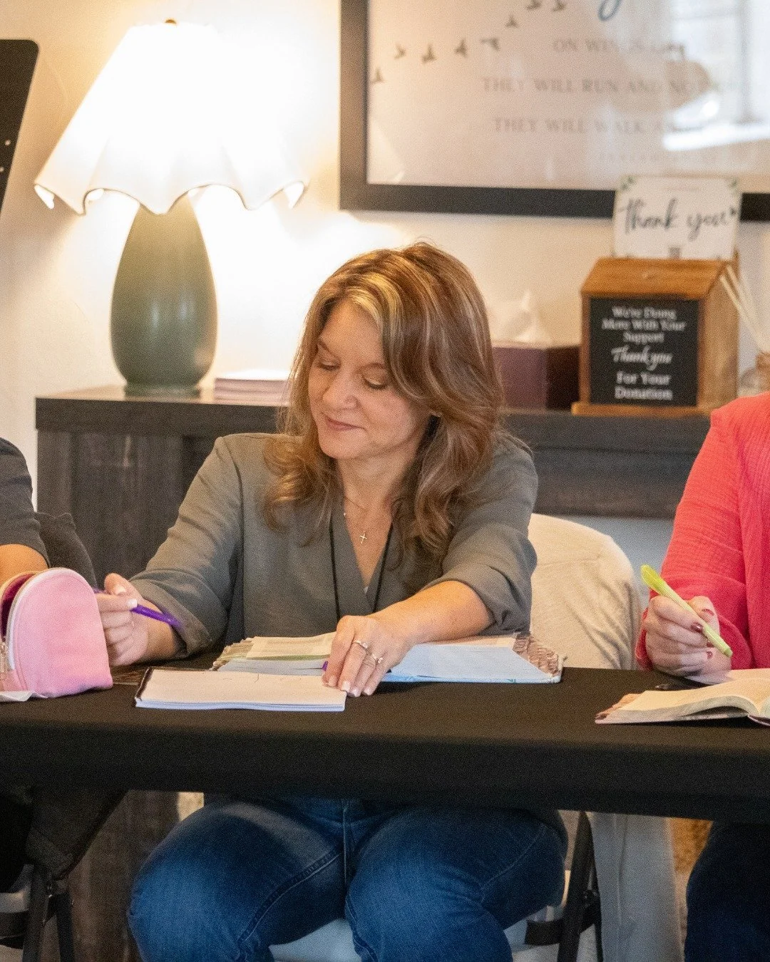 A woman sitting at a table, writing in a notebook with a purple pen, in a cozy indoor setting with a lamp and framed wall art in the background.