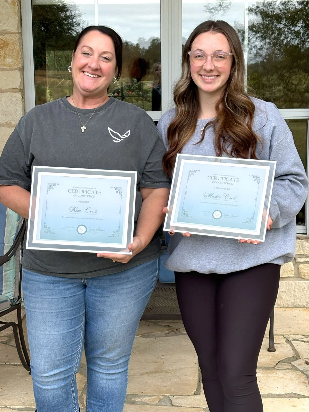 Two women standing outside in front of a window, each holding a certificate of completion and smiling at the camera.