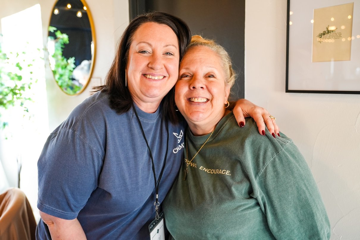 Two women hugging and smiling inside a well-lit room.