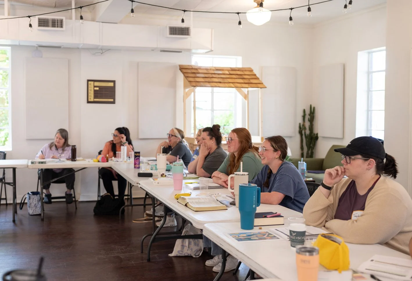 Women sitting at a long conference table in a bright room, smiling and listening, with notebooks and travel mugs in front of them, during a workshop or meeting.