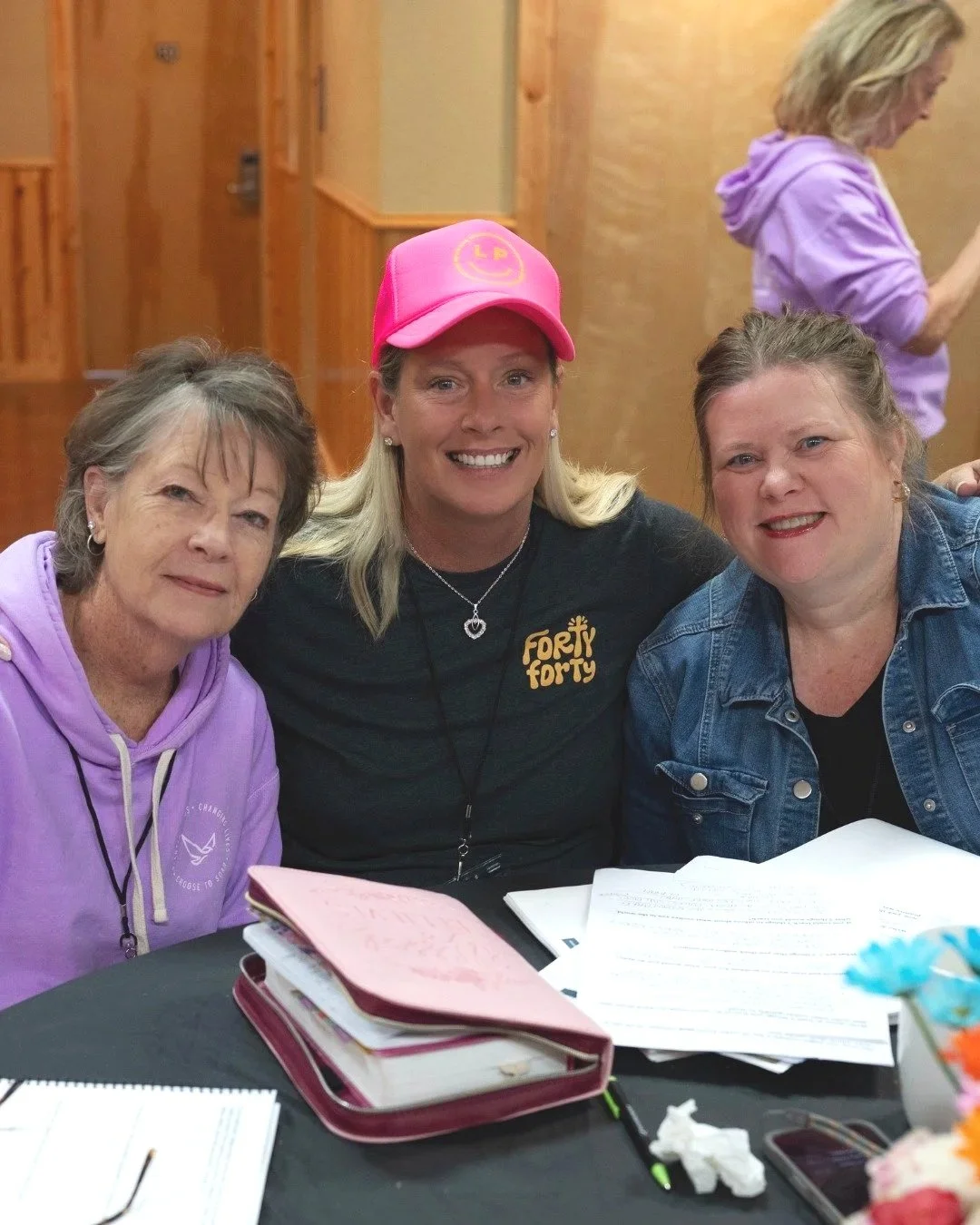 Three women sitting at a table, smiling at the camera. The woman in the middle wears a pink cap and a black shirt with "Forty Forty" logo, with papers and notebooks on the table.
