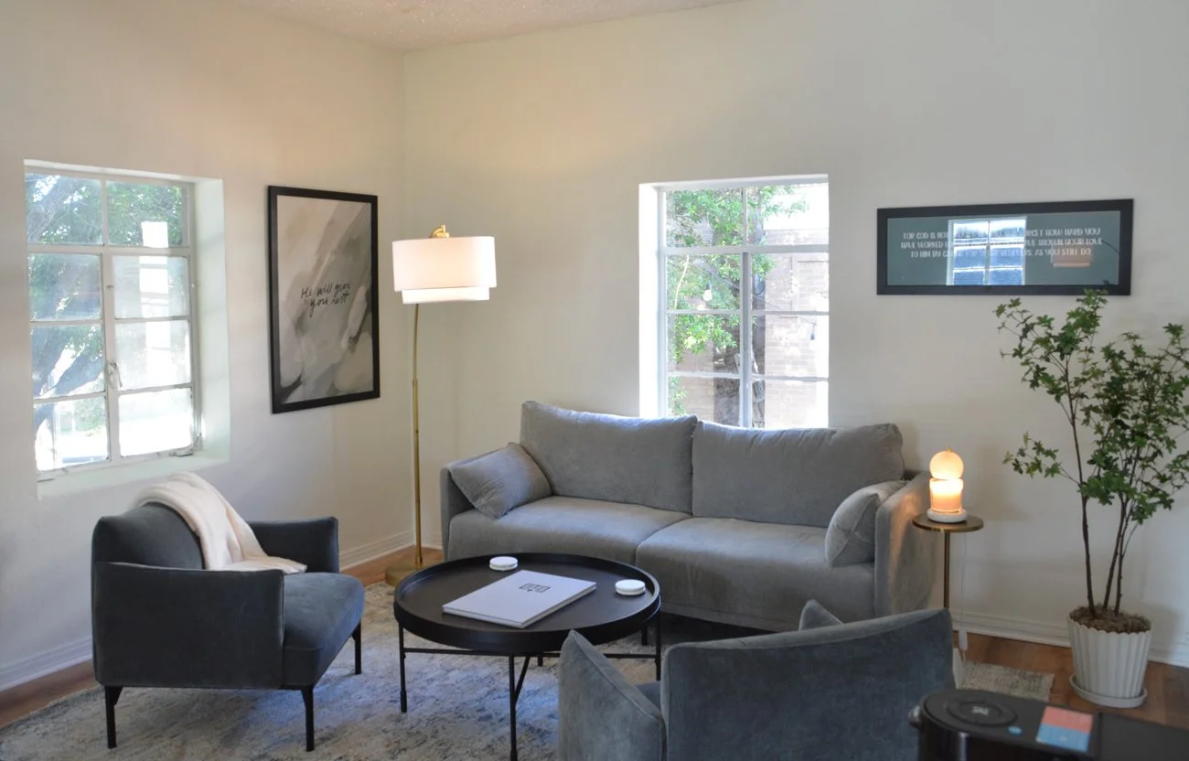 A cozy living room with a gray couch, a black armchair with a light blanket, a small round black coffee table, a tall floor lamp with a white lampshade, a potted plant, and framed wall art, with natural light coming through the windows.