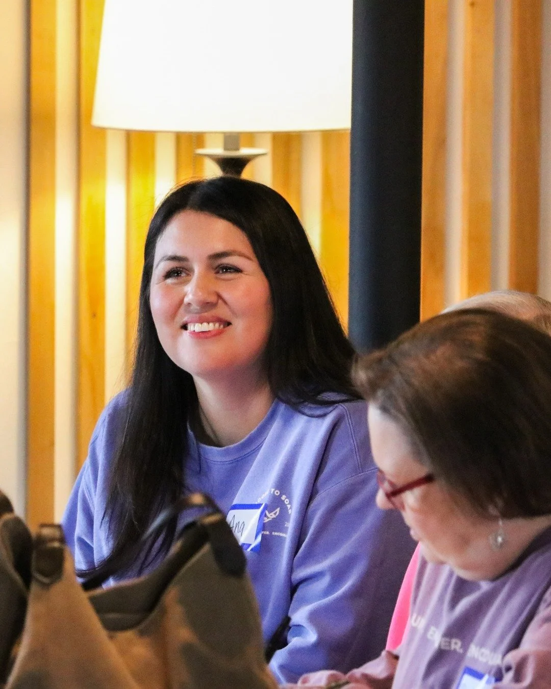 A woman with long dark hair smiling during an indoor event, wearing a purple shirt, with a lamp and wooden wall in the background.