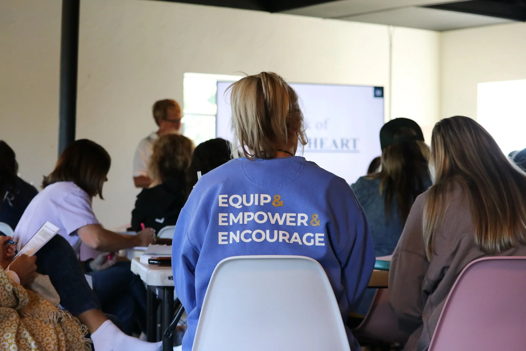 A group of people attending a seminar or workshop in a room with a presentation screen at the front. The focus is on a person wearing a blue sweatshirt with the words 'EQUIP & EMPOWER & ENCOURAGE' printed on the back, sitting among the audience.