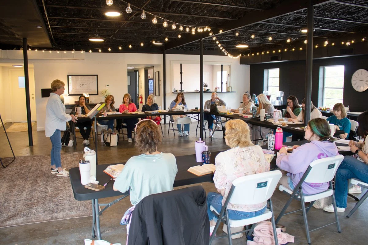 A group of women attending a workshop or class in a well-lit room, seated around U-shaped tables, with a woman standing and speaking at a podium.