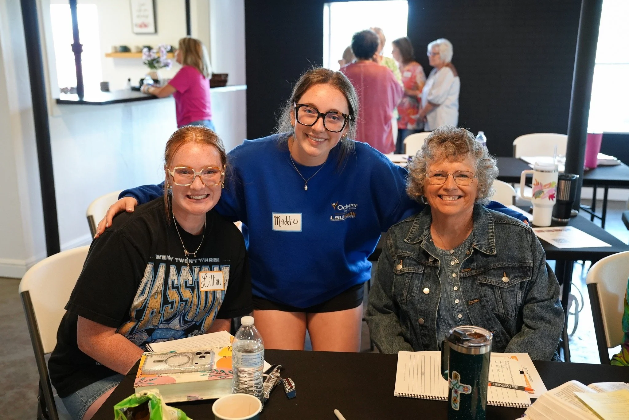 Three women sitting at a table and smiling at the camera, with a social gathering happening in the background in a bright room.