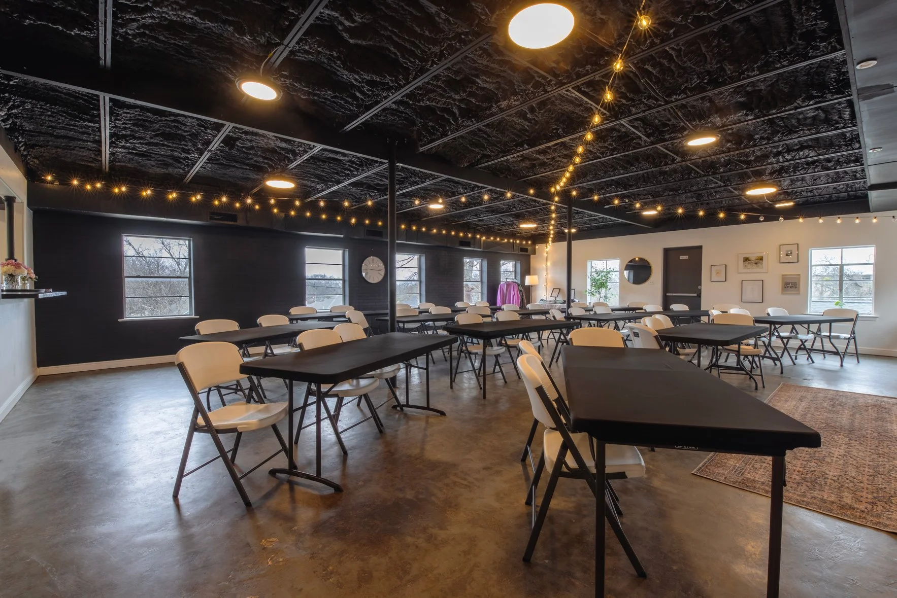A spacious event room with black ceiling, string lights, and large windows. Tables with black tablecloths and white chairs are arranged for a gathering. Decorative artwork and a round mirror on the white wall, with a rug near the entrance.