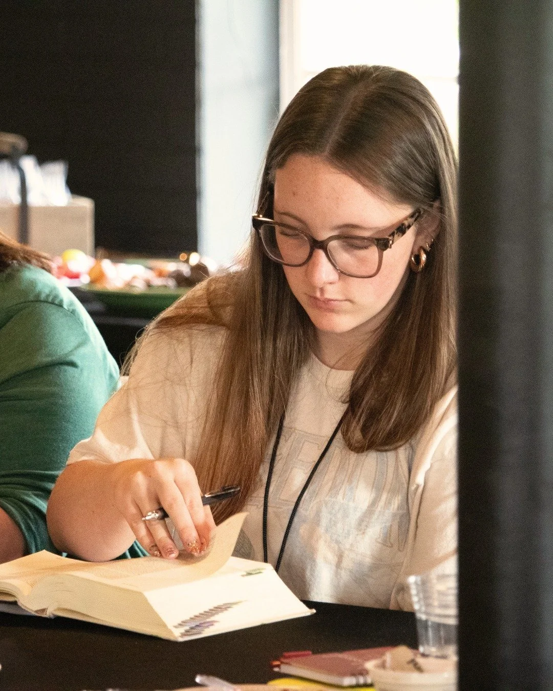 A young woman with long brown hair, glasses, and earrings, writing in a notebook at a table, with a black curtain and window in the background.