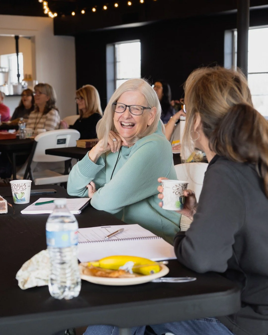 Women sitting at a table, smiling and talking, with notebooks, paper cups, and snacks on the table, during a group meeting or workshop.
