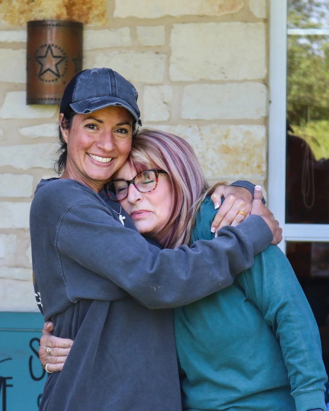 Two women are hugging outside a house with a stone wall. One woman is wearing a black cap and gray sweatshirt, and the other woman is wearing glasses and a teal shirt.