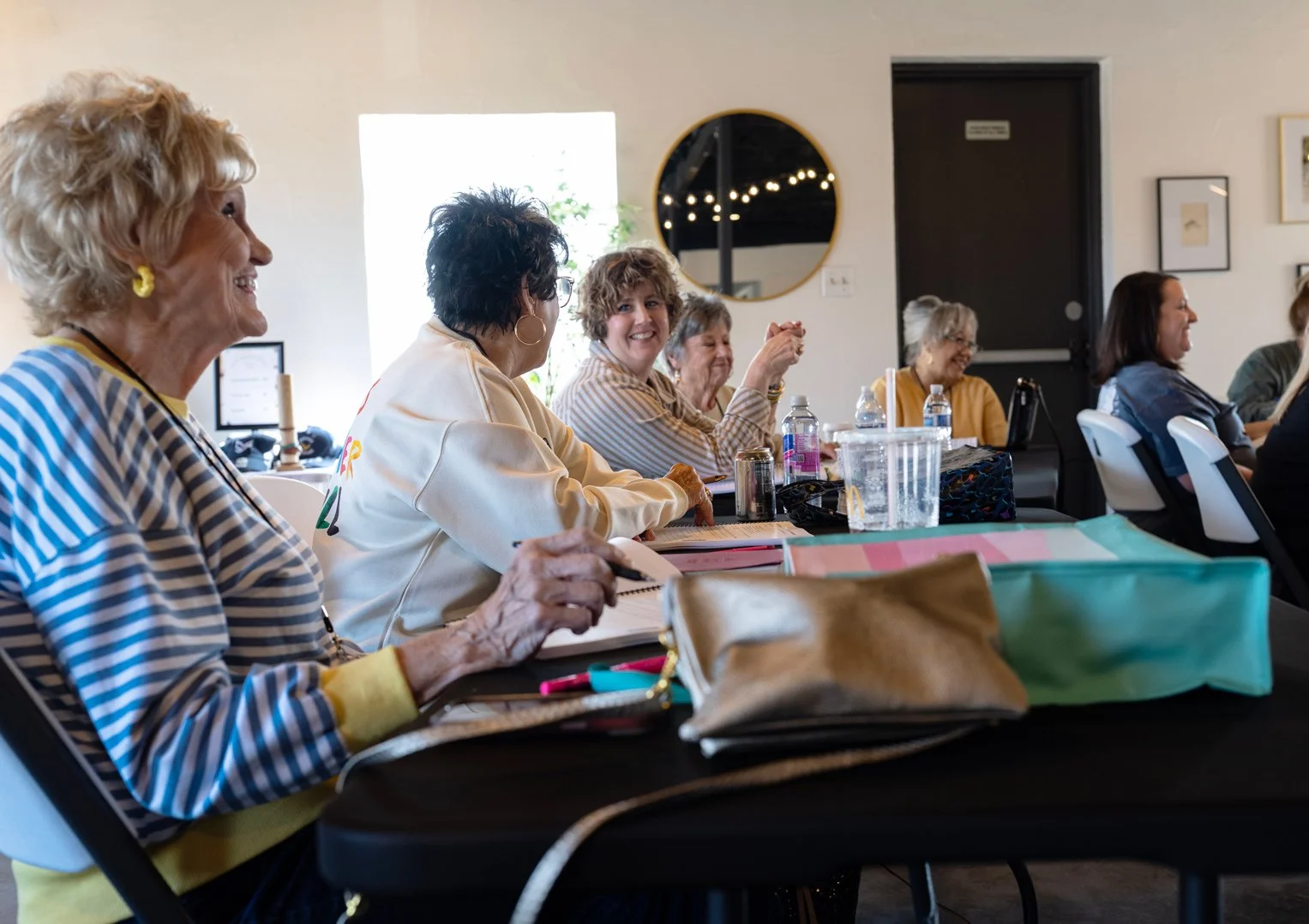 A group of women sitting at a table, smiling and engaging in conversation during a gathering in a well-lit room with framed artwork on the walls.