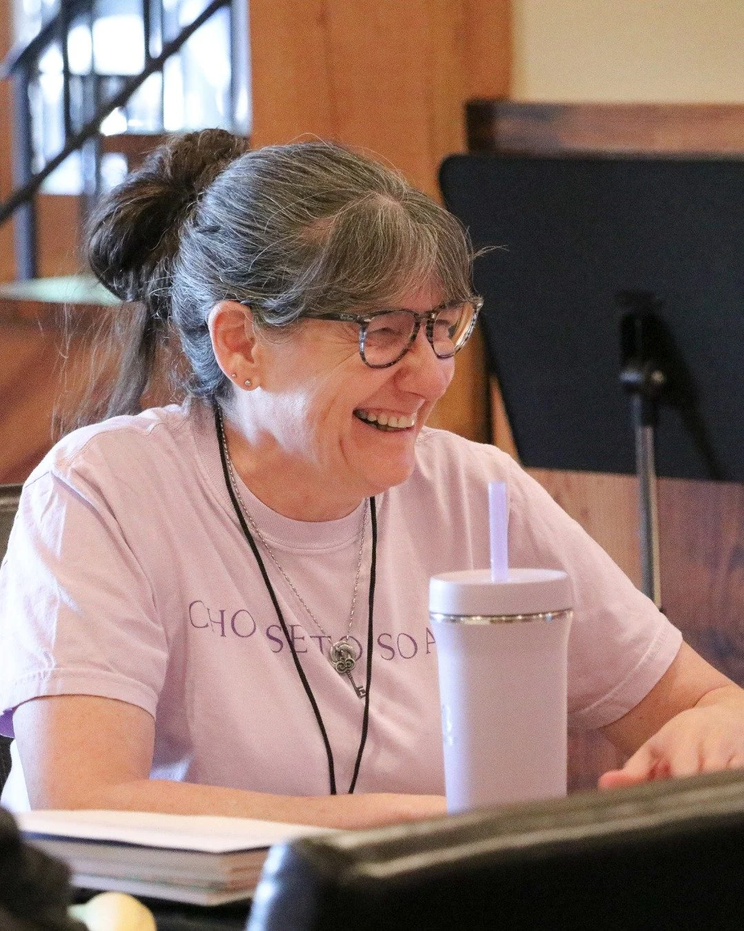 Woman with glasses and gray hair in a ponytail, smiling and laughing, sitting at a table with a white tumbler and a notepad, in a cozy indoor setting.