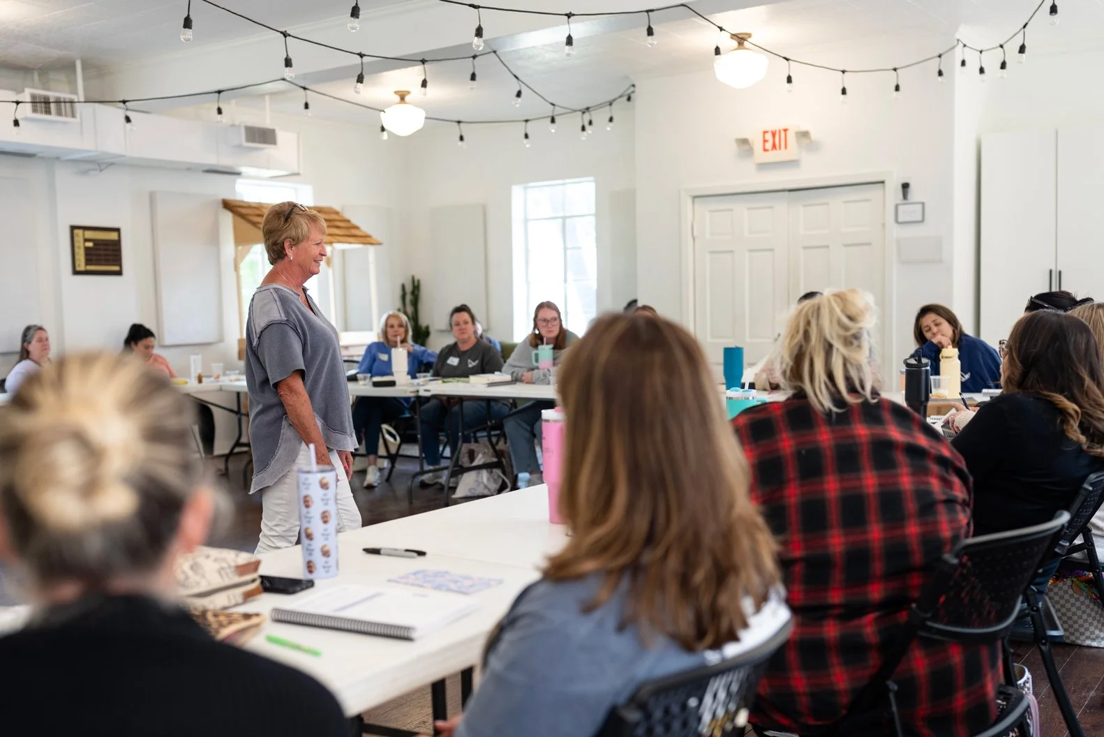 A woman leading a workshop or seminar with attendees seated around tables in a bright, decorated room.