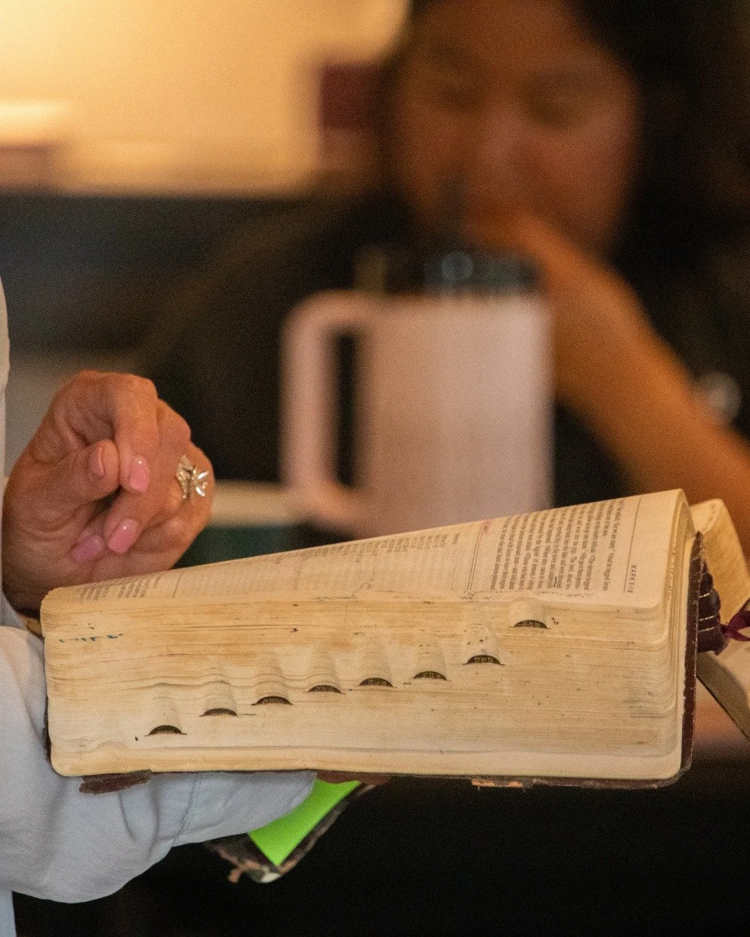 Close-up of a person holding an open, worn Bible with visible bookmark tabs. In the background, a woman with long curly hair is taking a photo with her phone, with a blurred microwave oven behind her.