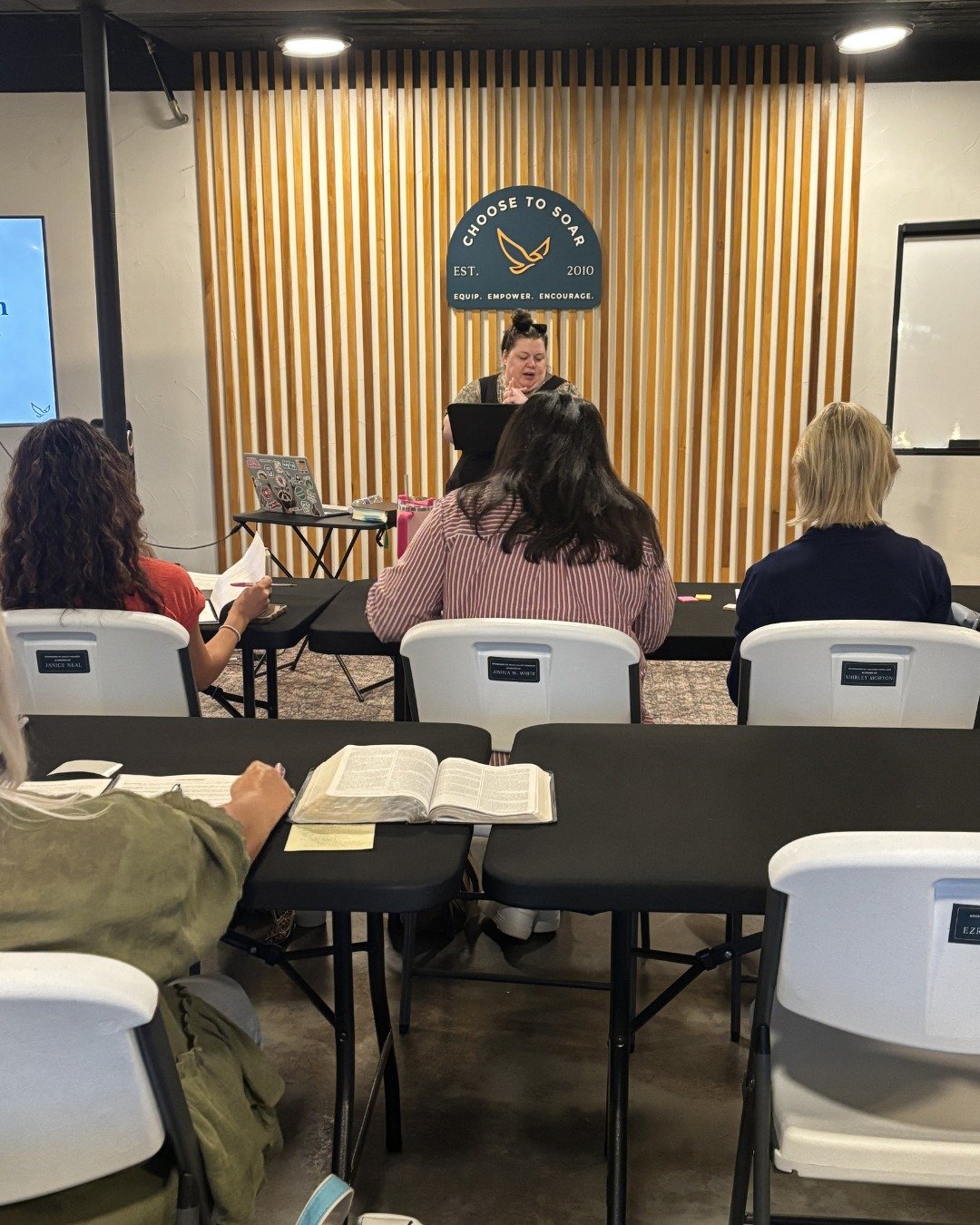 A woman standing at a podium giving a presentation to an audience sitting at tables in a room with wooden slats on the wall behind her. There is a sign that reads 'Choose to Soar' and an electronic screen on the side.