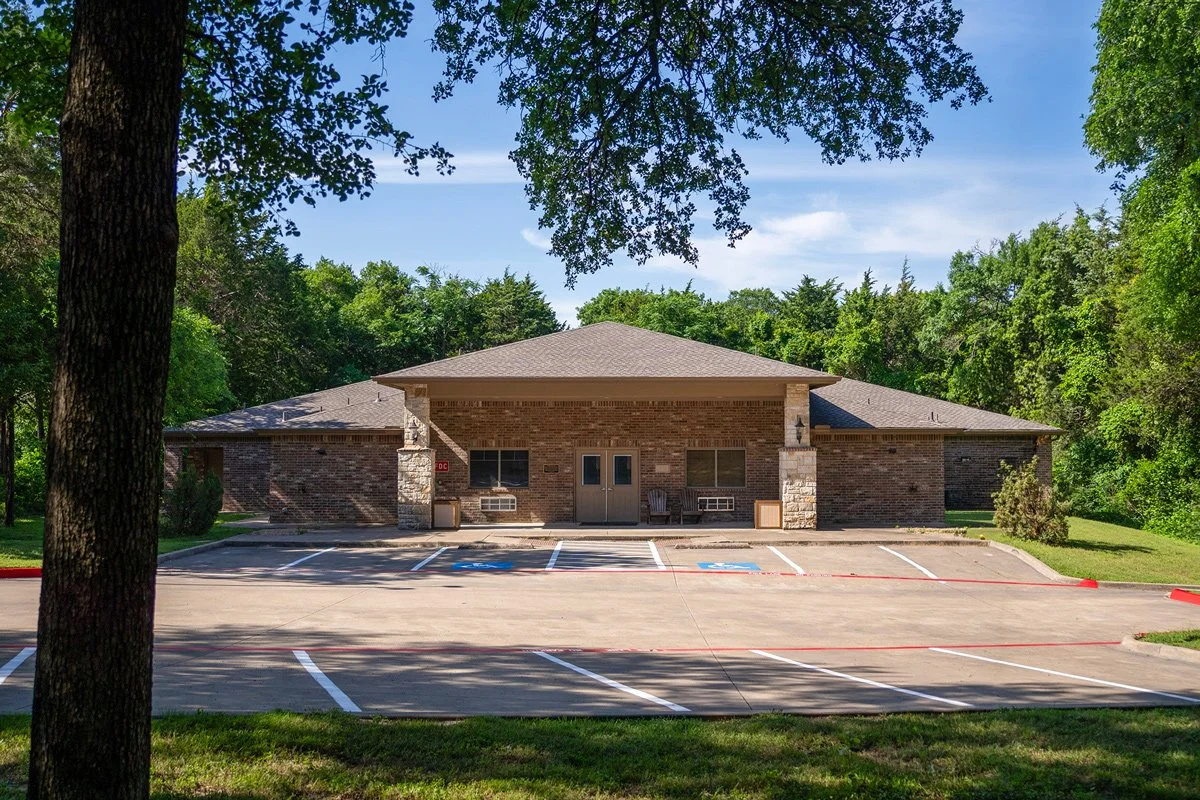 A single-story brick building with a parking lot in front, including designated handicapped parking spaces, surrounded by green trees and grass under a blue sky.