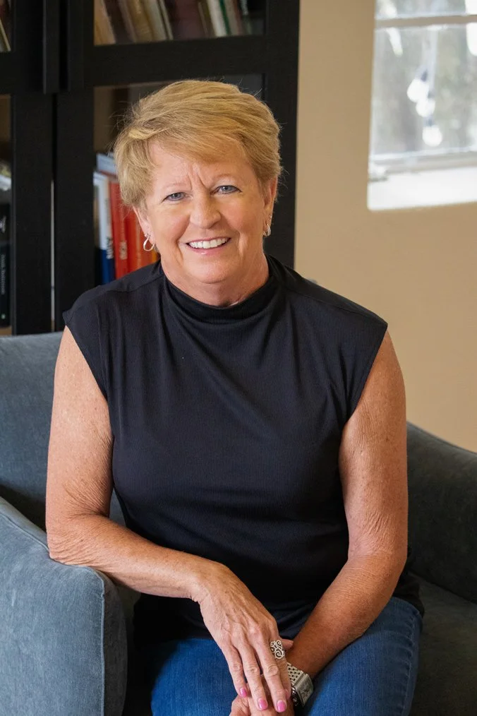 A woman with short blonde hair, wearing a black sleeveless top, sitting on a gray couch in a room with a black bookshelf and a window in the background.
