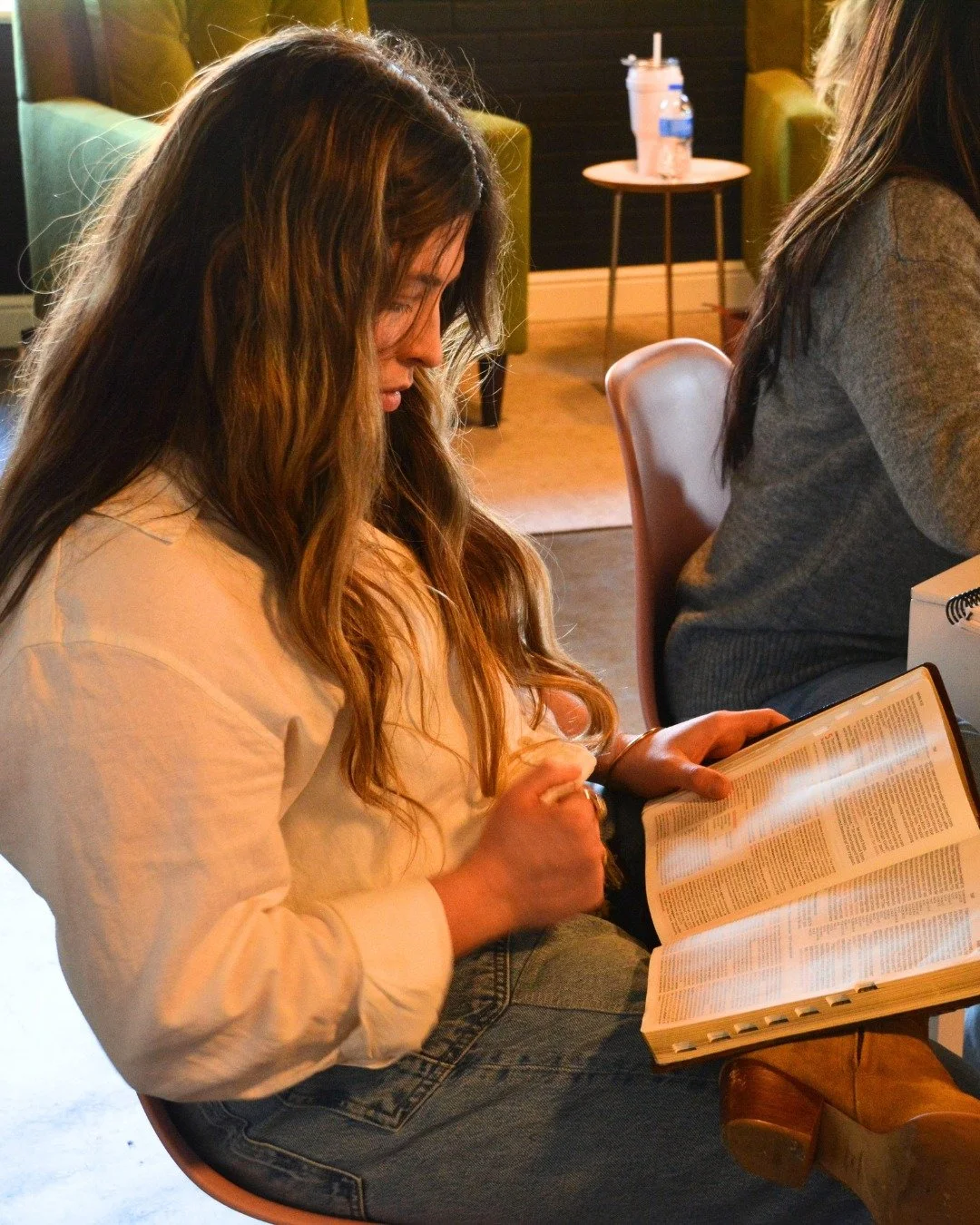 A woman with long wavy hair, wearing a cream-colored jacket and jeans, looking down at a book she is holding, sitting indoors with warm lighting.