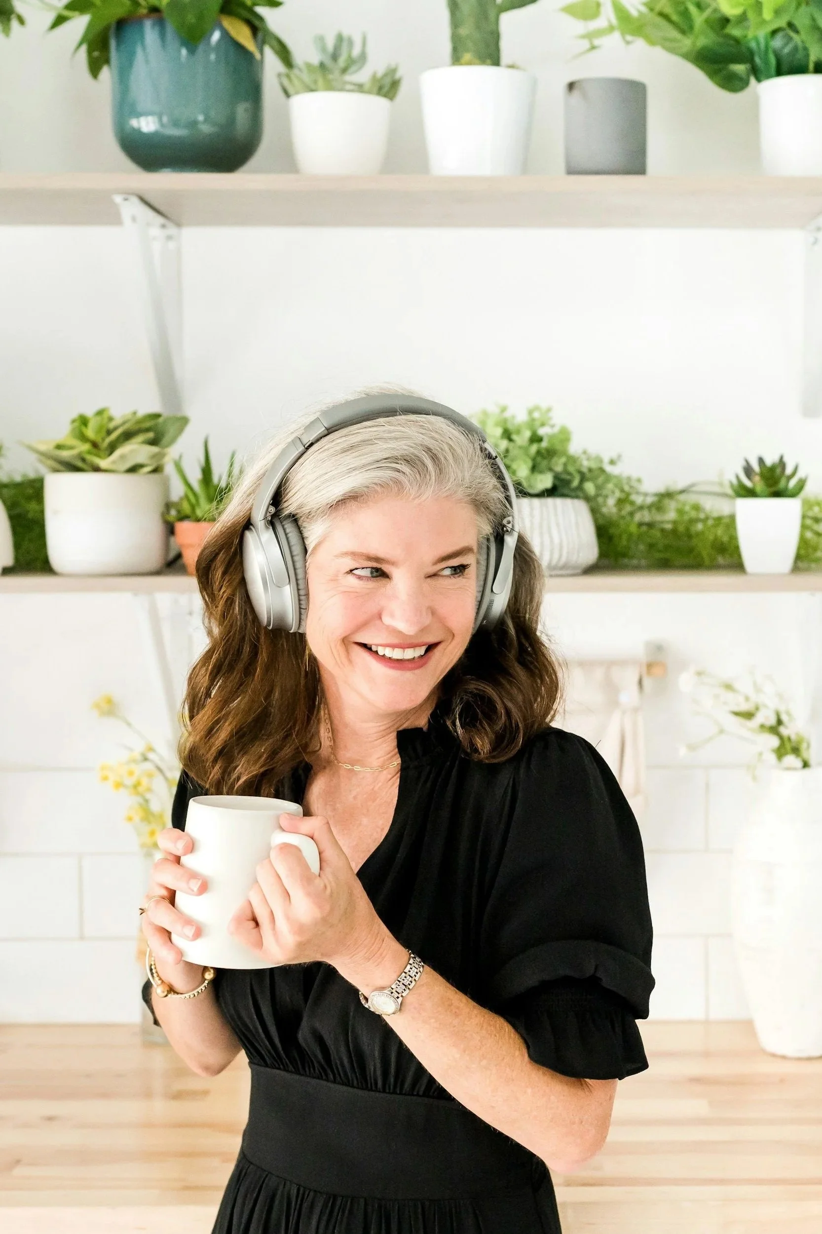 A woman with gray hair wearing a black dress is smiling and holding a white mug with both hands. She is wearing headphones and looking to the side. Behind her, there are shelves with various potted plants.