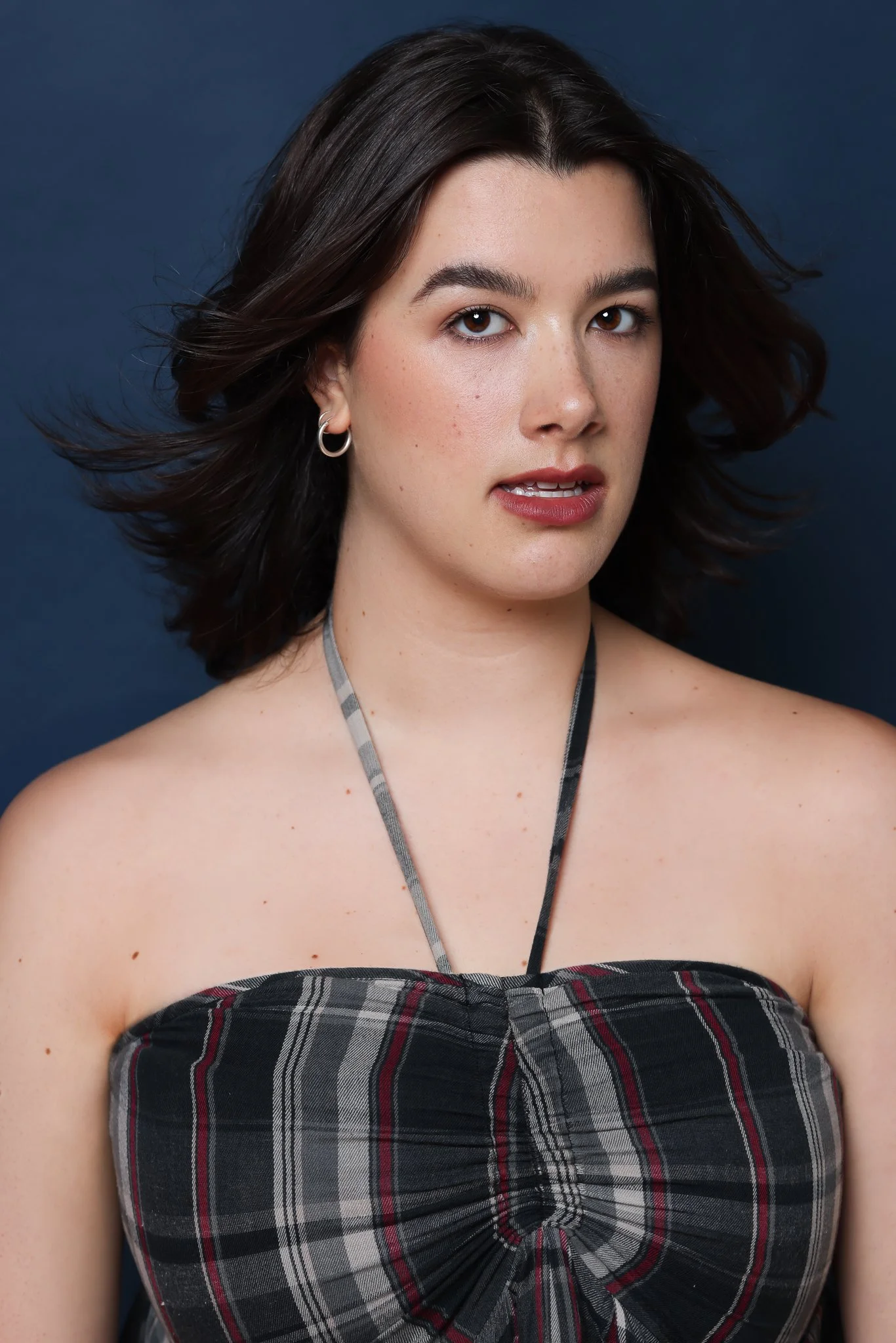 Close-up of a young woman with short dark hair, wearing a plaid strapless top with a gray and red pattern, standing against a dark blue background.