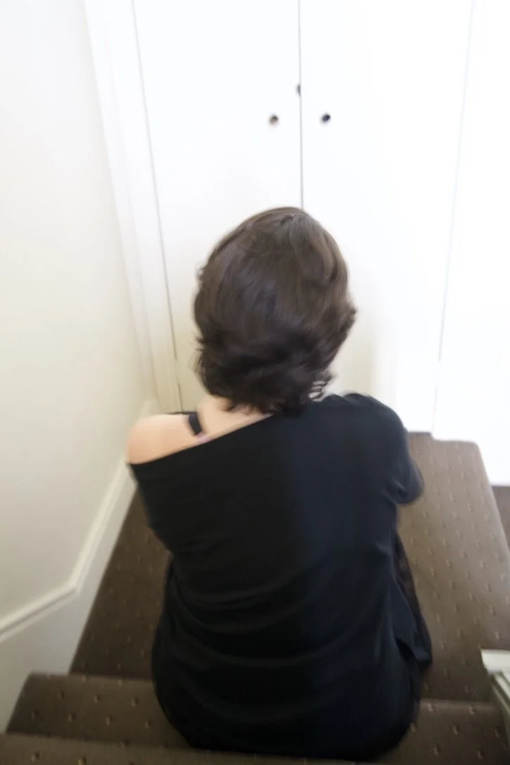 A person with short, wavy brown hair sitting on a carpeted staircase, facing away, wearing a black shirt with one shoulder exposed.
