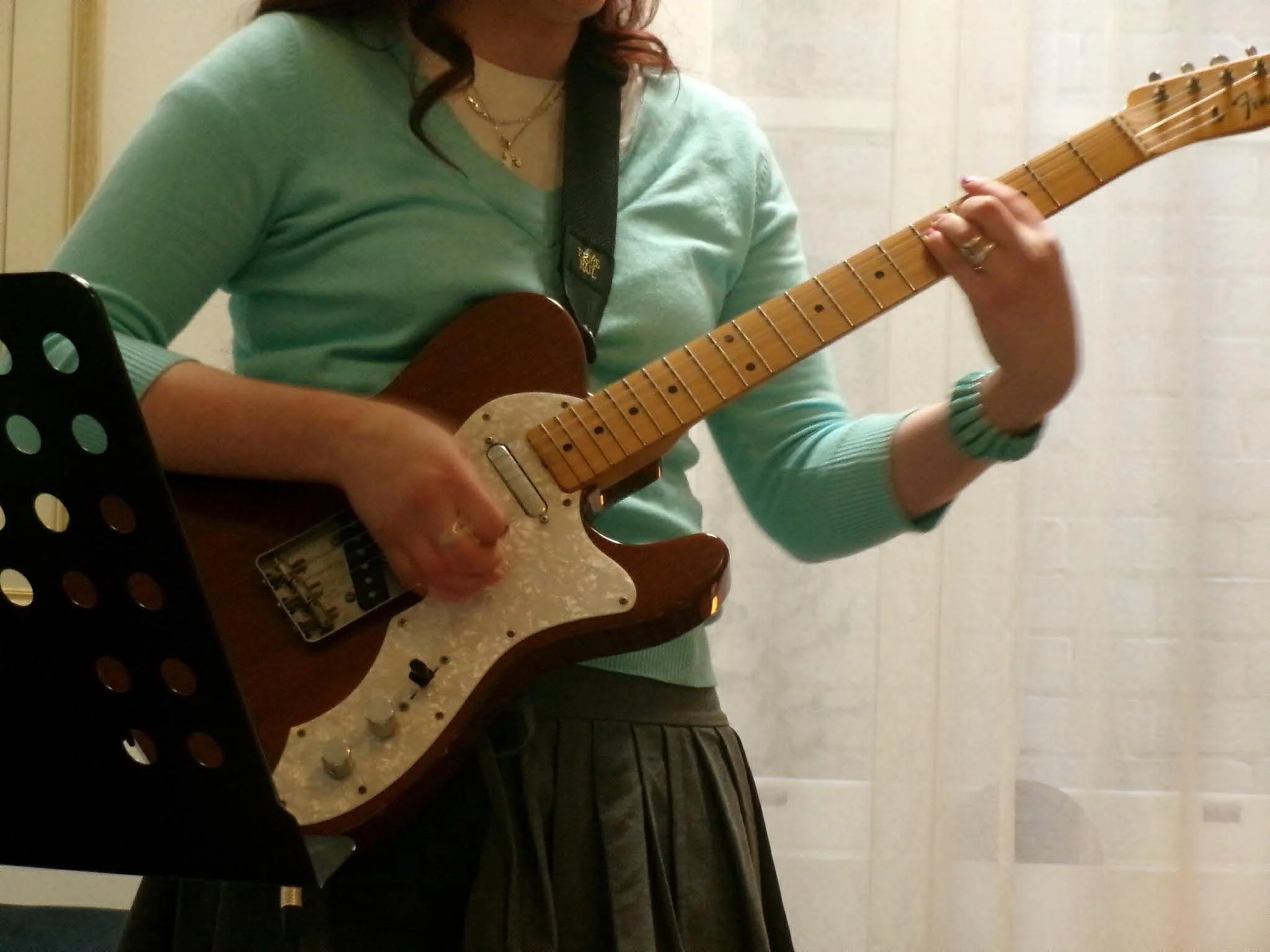 Person playing an electric guitar, wearing a light green long-sleeve shirt, gold necklaces, and a bracelet, with a music stand nearby, in an indoor setting with light-colored tiled walls.