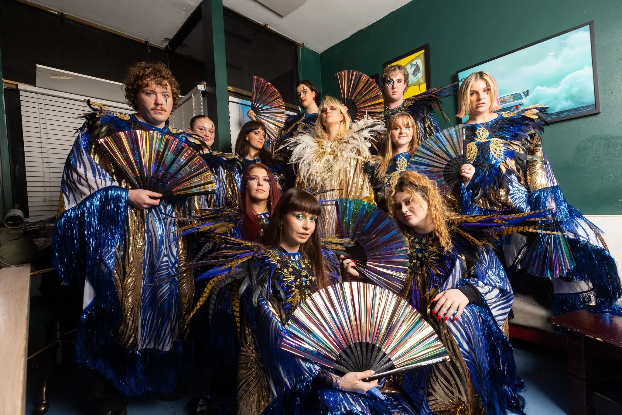 Group of performers in elaborate costumes with feathers and fans inside a room.