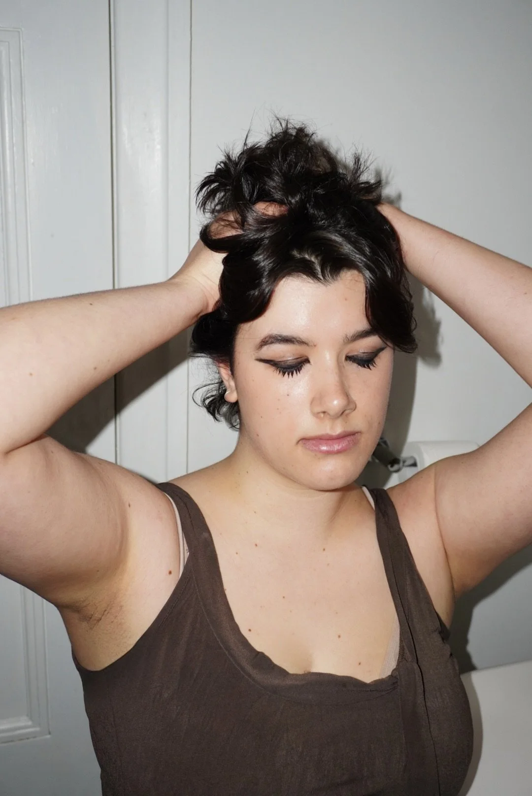 A young woman with short, dark, wavy hair, wearing a brown tank top, with her eyes closed and hands in her hair, standing in front of a white door.