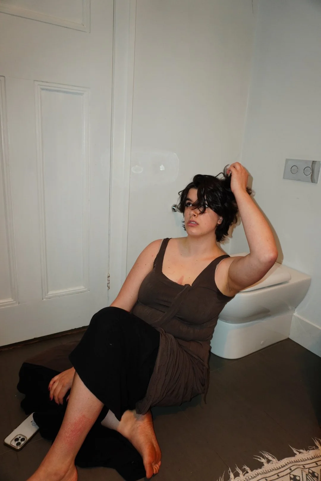 A woman with short dark hair sitting on the floor in front of a white door, leaning against a wall with her hand in her hair, next to a wall-mounted toilet.