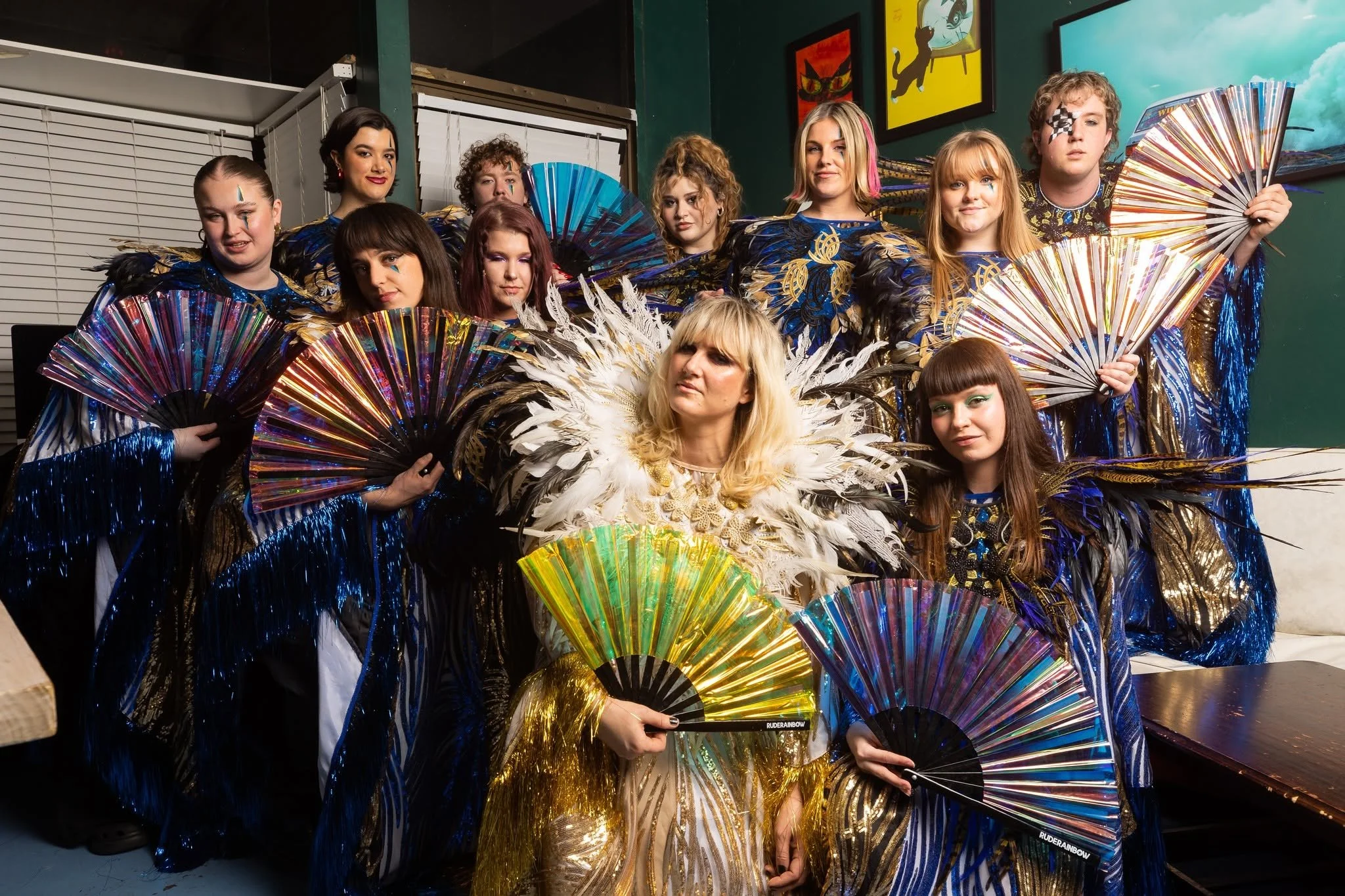 Group of people dressed in elaborate costumes with feathers and holding colorful fans, posing together indoors.