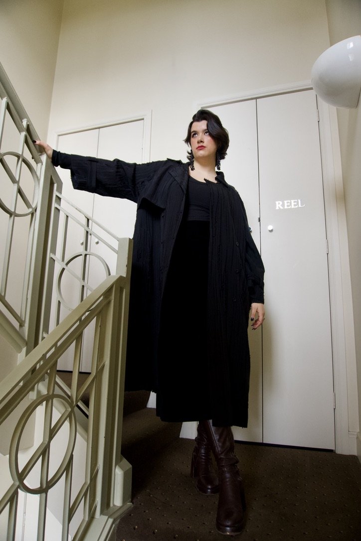 A woman with dark hair and light skin, dressed in all black, standing at the top of a staircase in an indoor setting, holding onto the railing with one arm, with a white door labeled 'REEL' behind her.