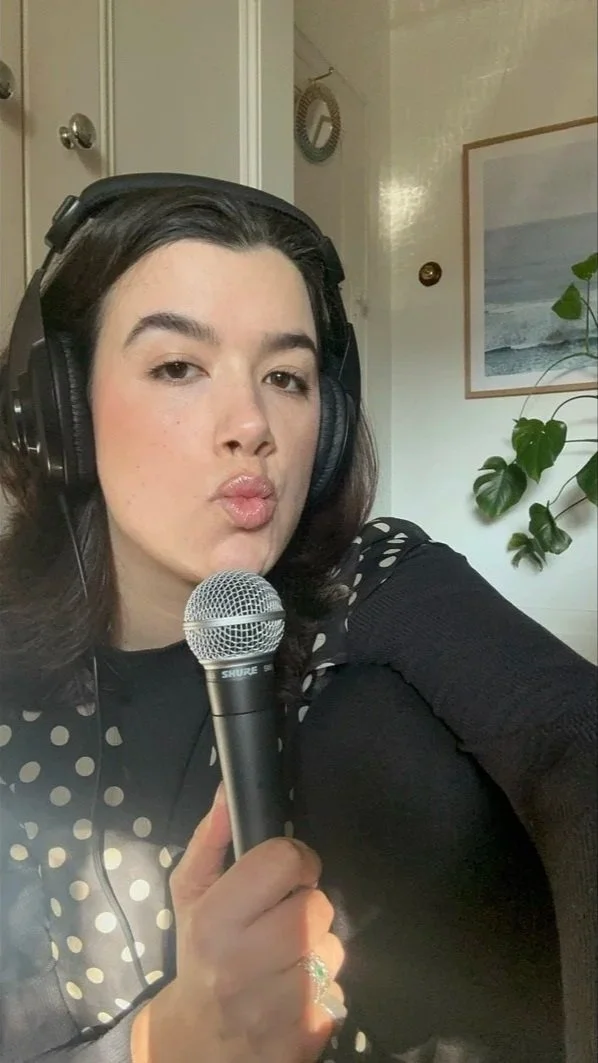 A woman with dark hair wearing large over-ear headphones, holding a microphone, and making a duck face expression. She is wearing a black top with polka-dot details on the shoulders, and a ring on her finger. There is a picture frame and a potted plant visible in the background.