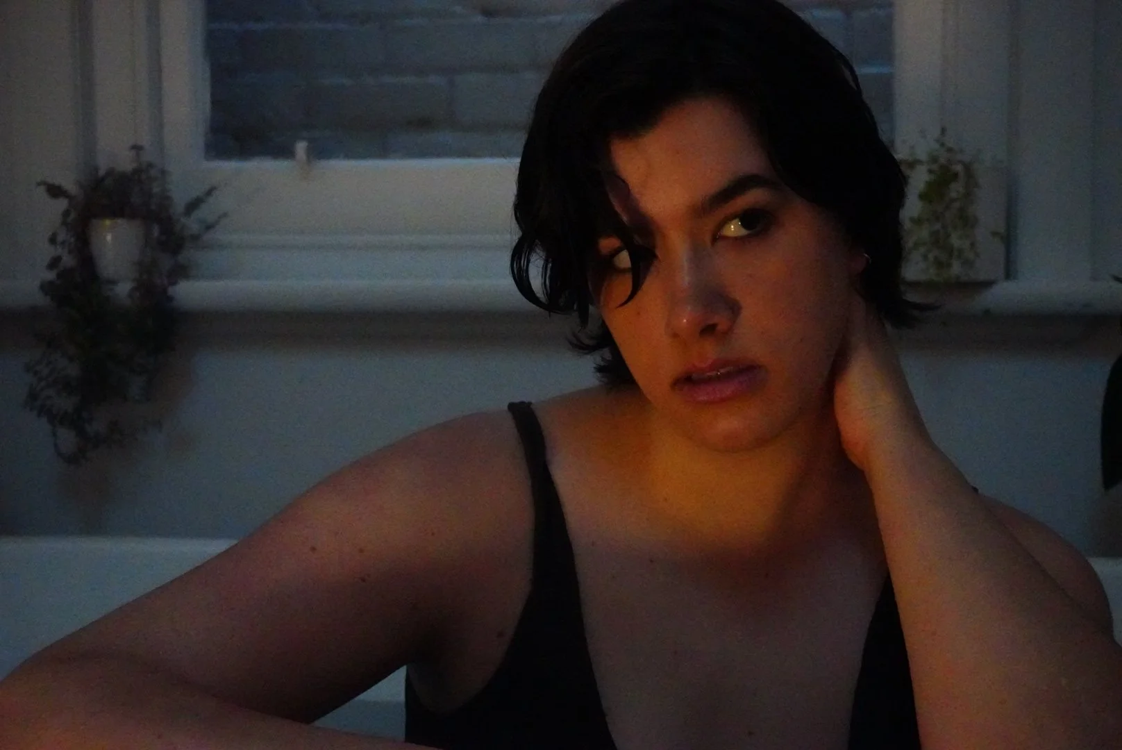 A young woman with short dark hair, wearing a black tank top, posing indoors near a window with blinds. She is touching her neck with one hand, looking directly at the camera with a neutral expression. The room has some plants on the windowsill.