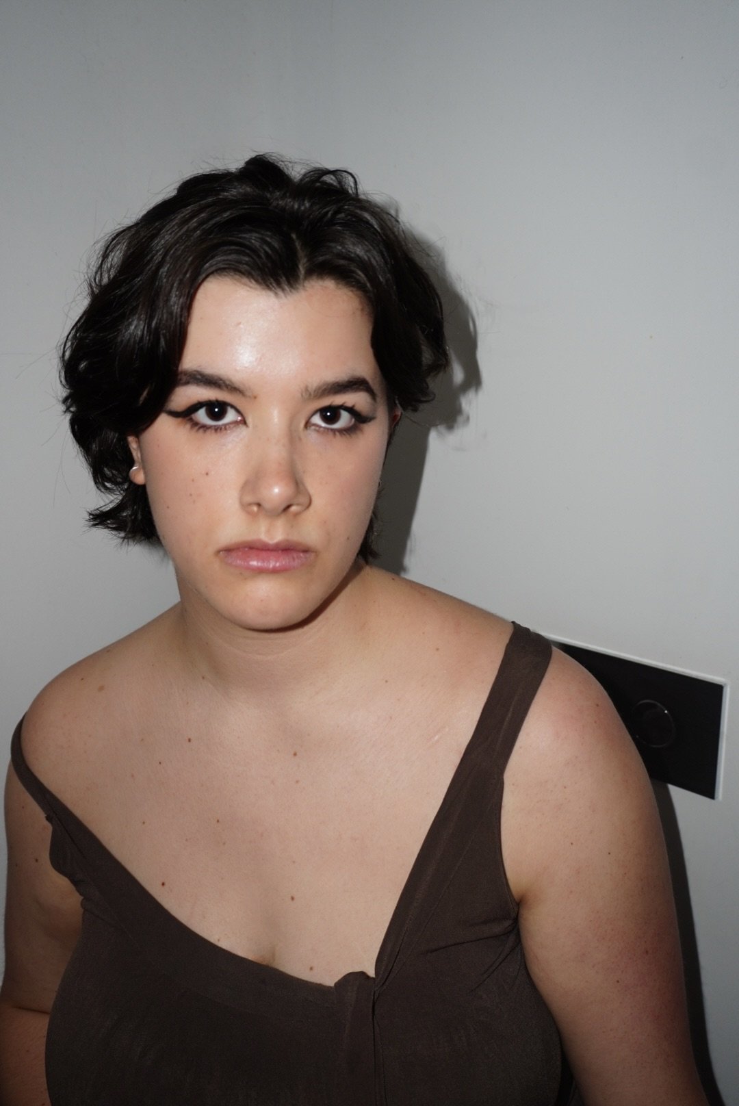 A young woman with short dark hair, wearing a brown sleeveless top, looking directly at the camera with a serious expression, standing against a plain light gray wall.