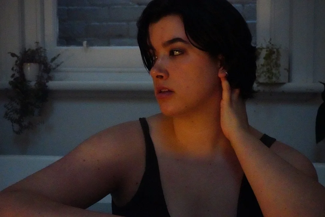 A woman with short dark hair touching her ear, sitting indoors near a window with plants on the windowsill, in low lighting.