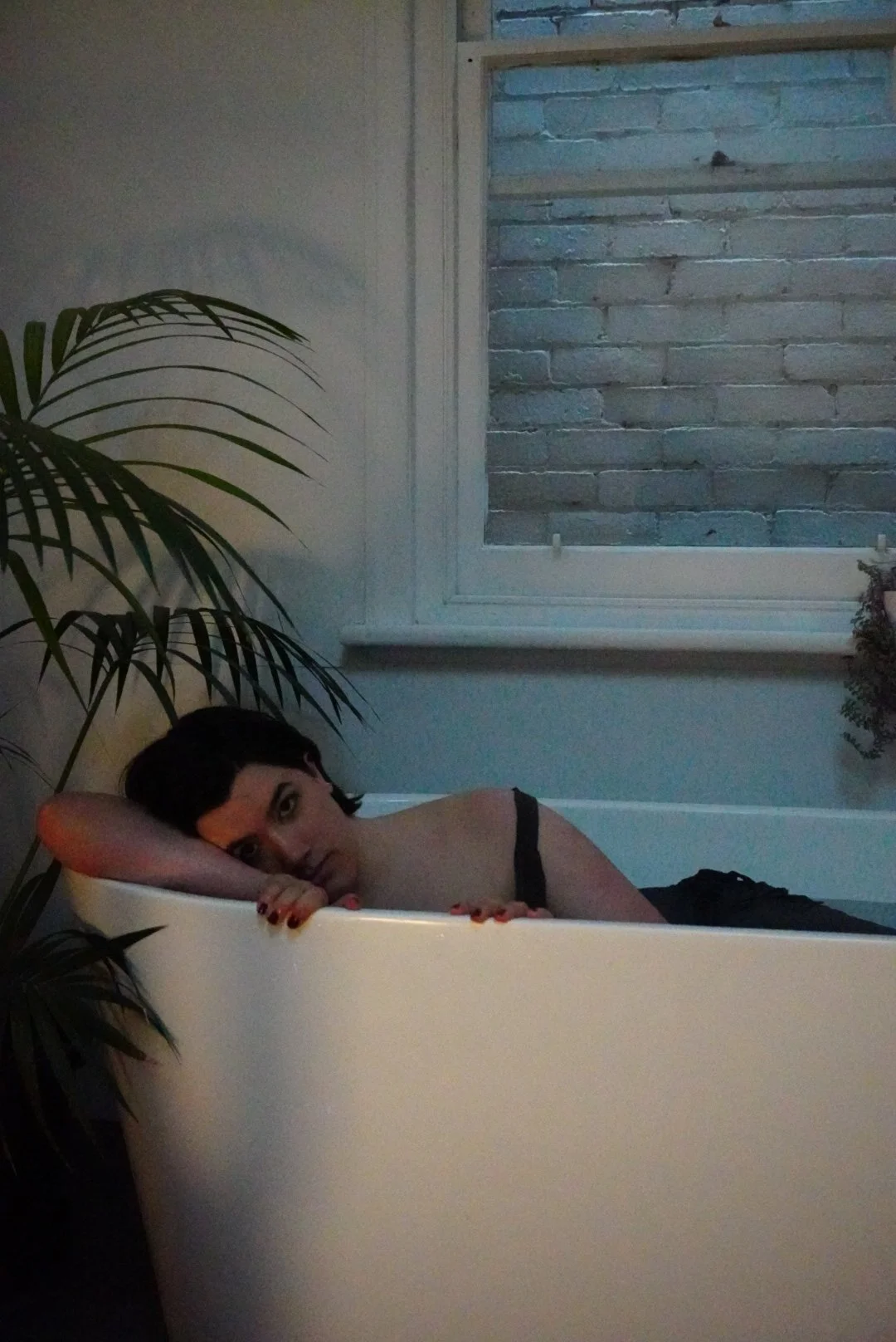 Woman with short black hair resting her head on her arm in a white bathtub next to a window with a brick wall outside, surrounded by indoor plants.