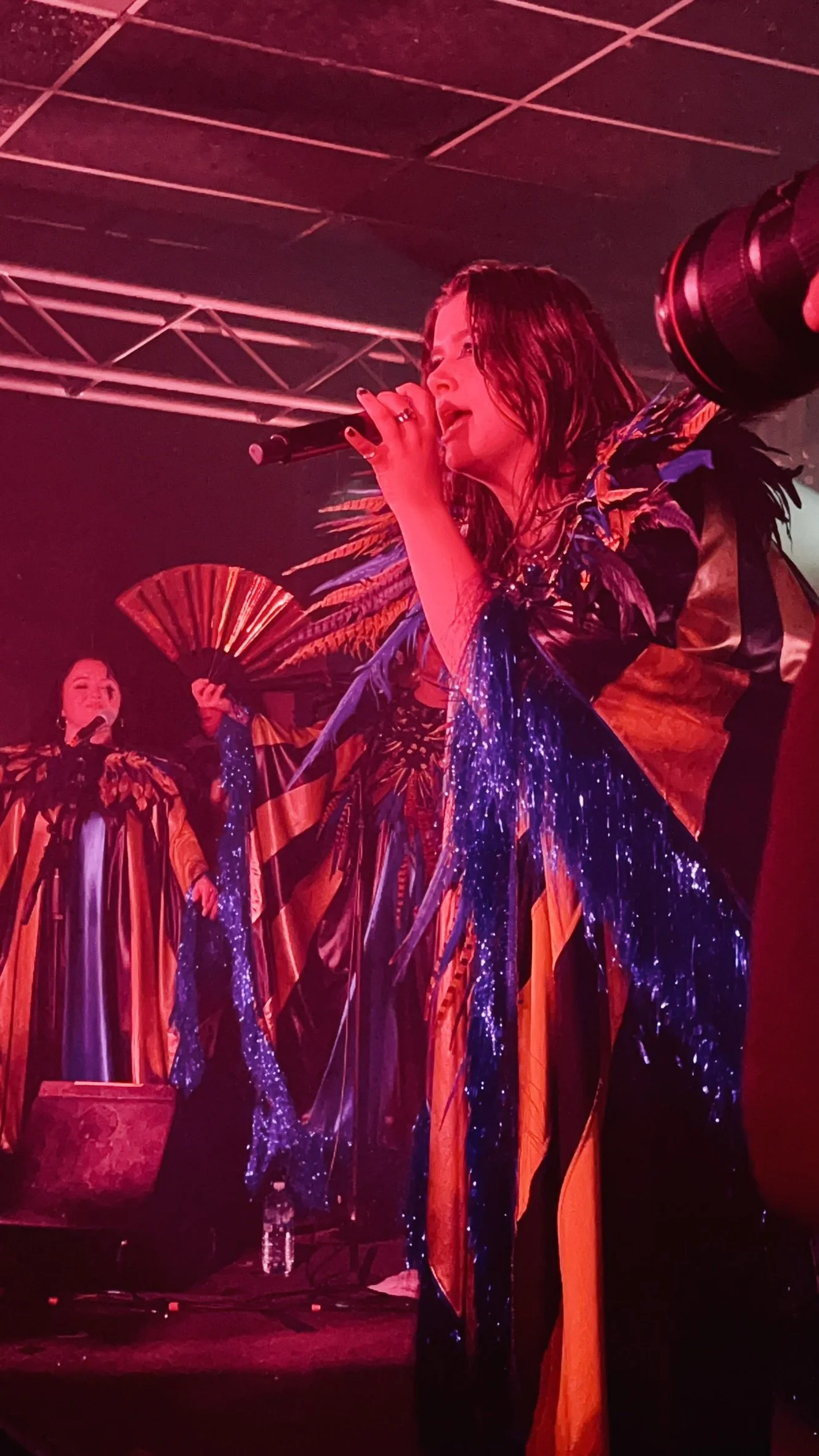 A performer singing into a microphone, dressed in a colorful, shiny costume with long flowing fabric, on stage with red lighting. There is another performer in the background holding a fan and wearing a similar ornate outfit.