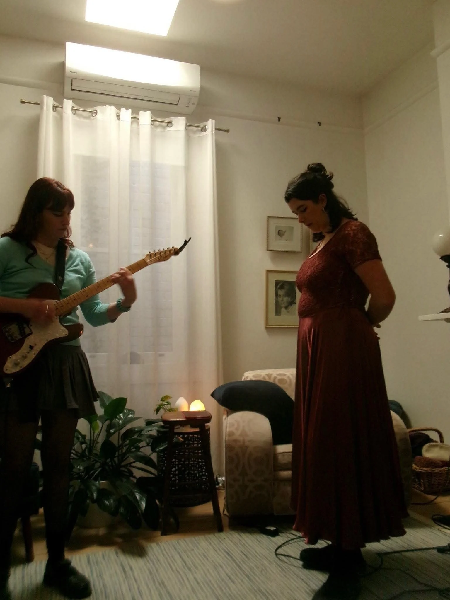 Two women are in a cozy living room, one playing an acoustic guitar while the other stands with her head bowed. The room has a patterned carpet, framed pictures on the wall, and plants near a window with white curtains. A small table with candles and a chair is also visible.