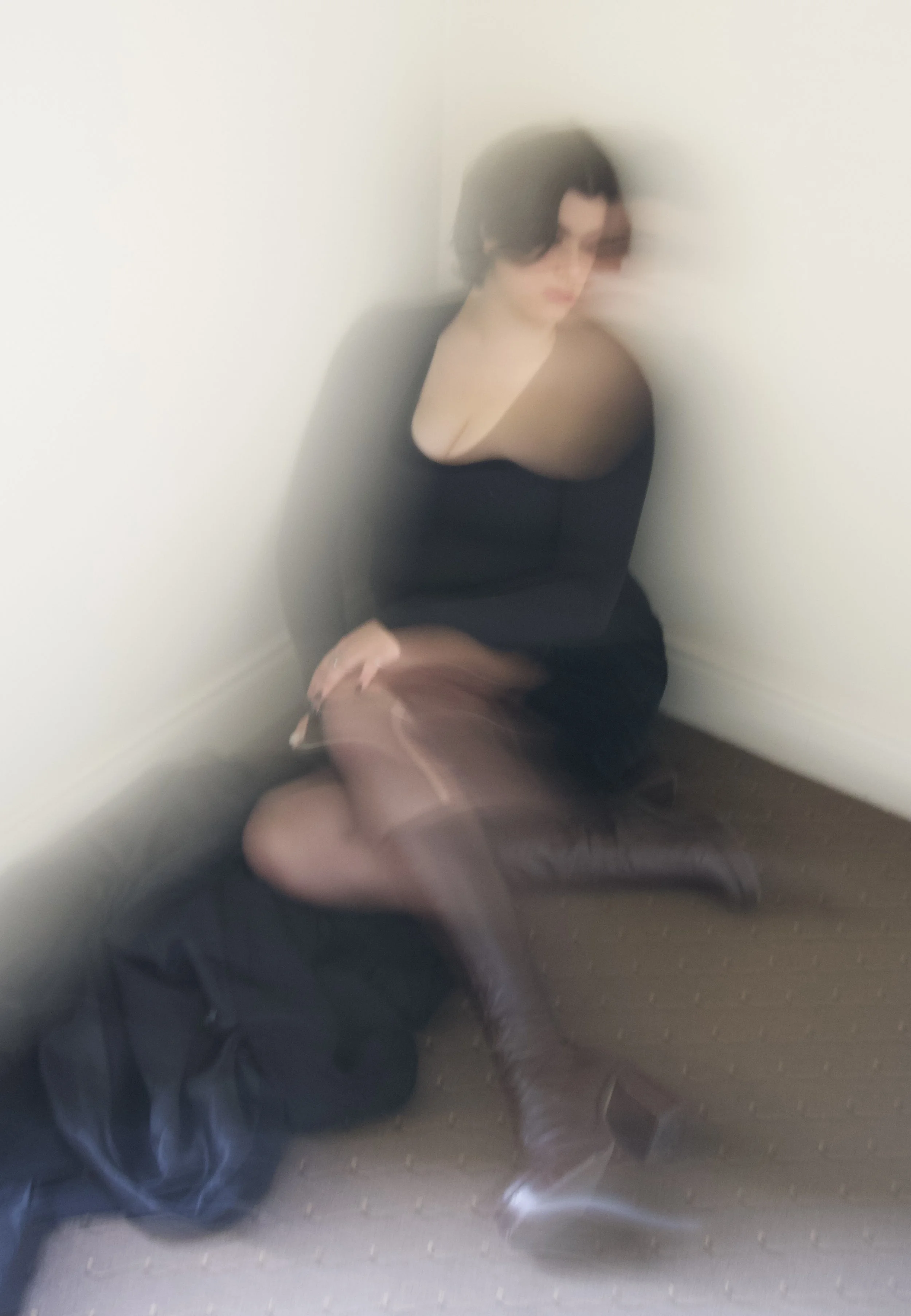 Blurred image of a woman with short dark hair leaning against a white wall, wearing a black top, and a man in dark clothing and boots standing nearby, with the scene slightly out of focus.