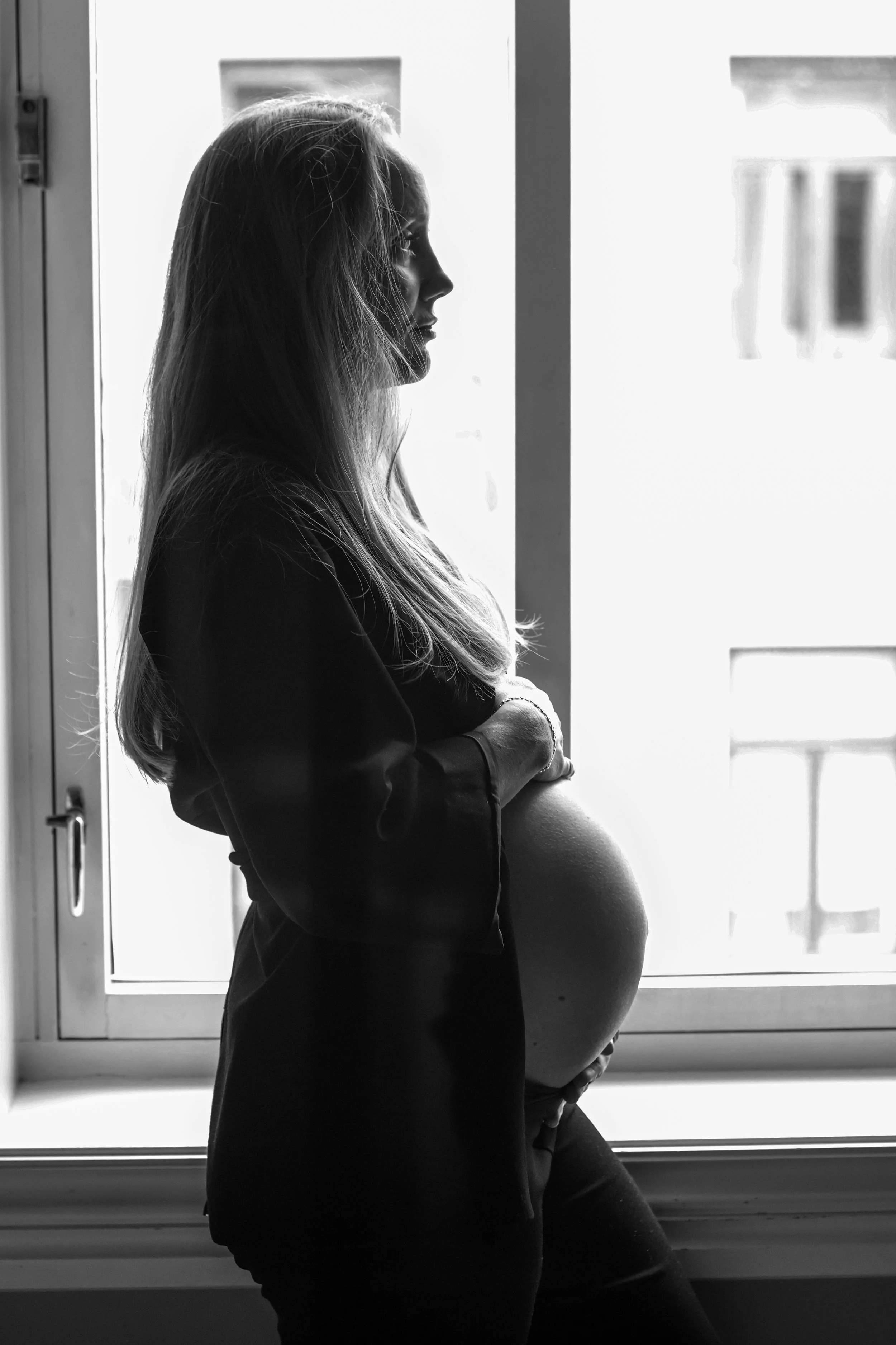 Black and white photo of a pregnant woman standing by a window, with her eyes closed and her arms crossed over her belly, in a peaceful indoor setting.