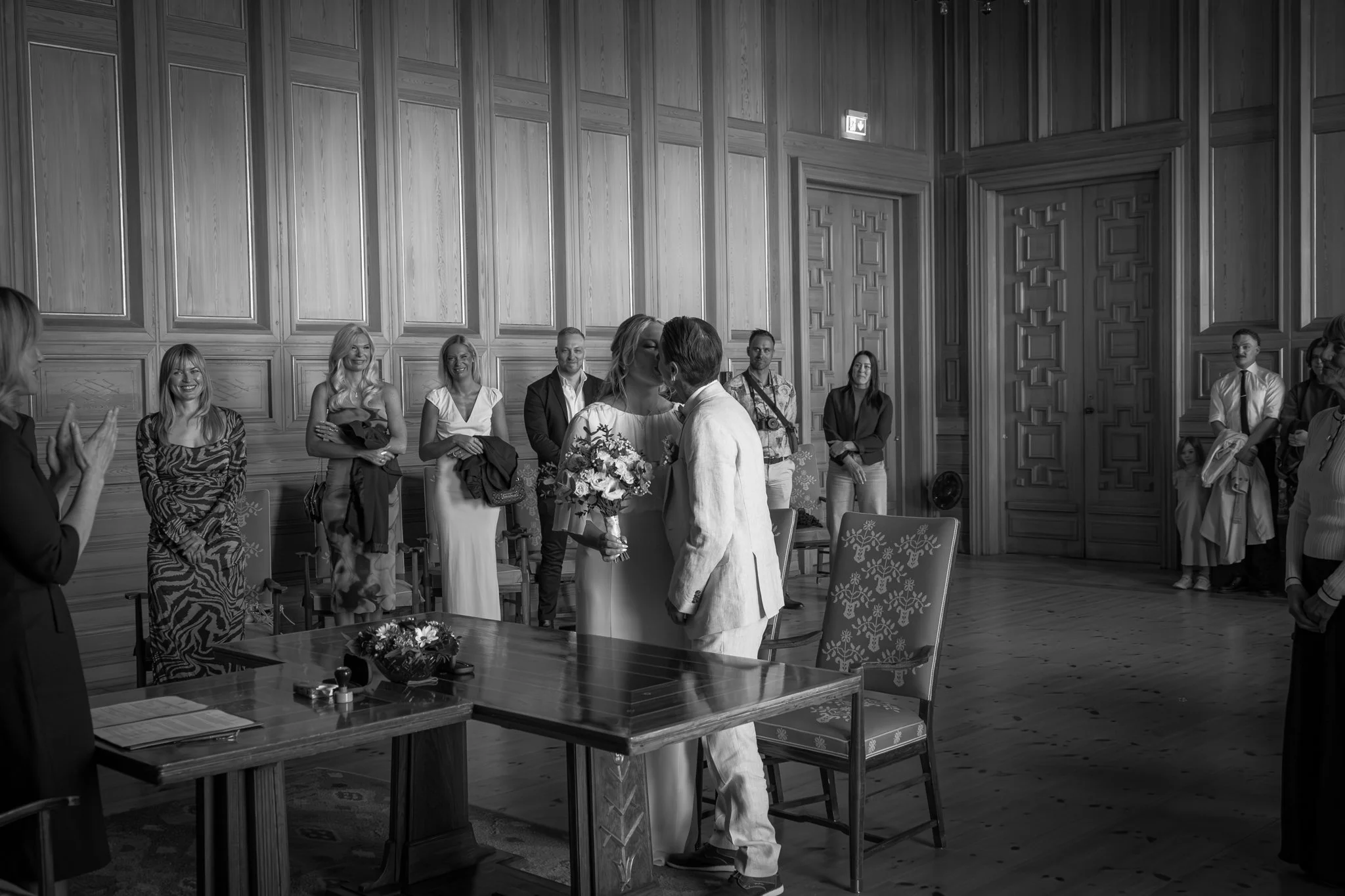 A wedding ceremony inside a large wooden hall with ornate wall paneling. A couple is exchanging vows, surrounded by friends and family, with some people clapping and smiling.