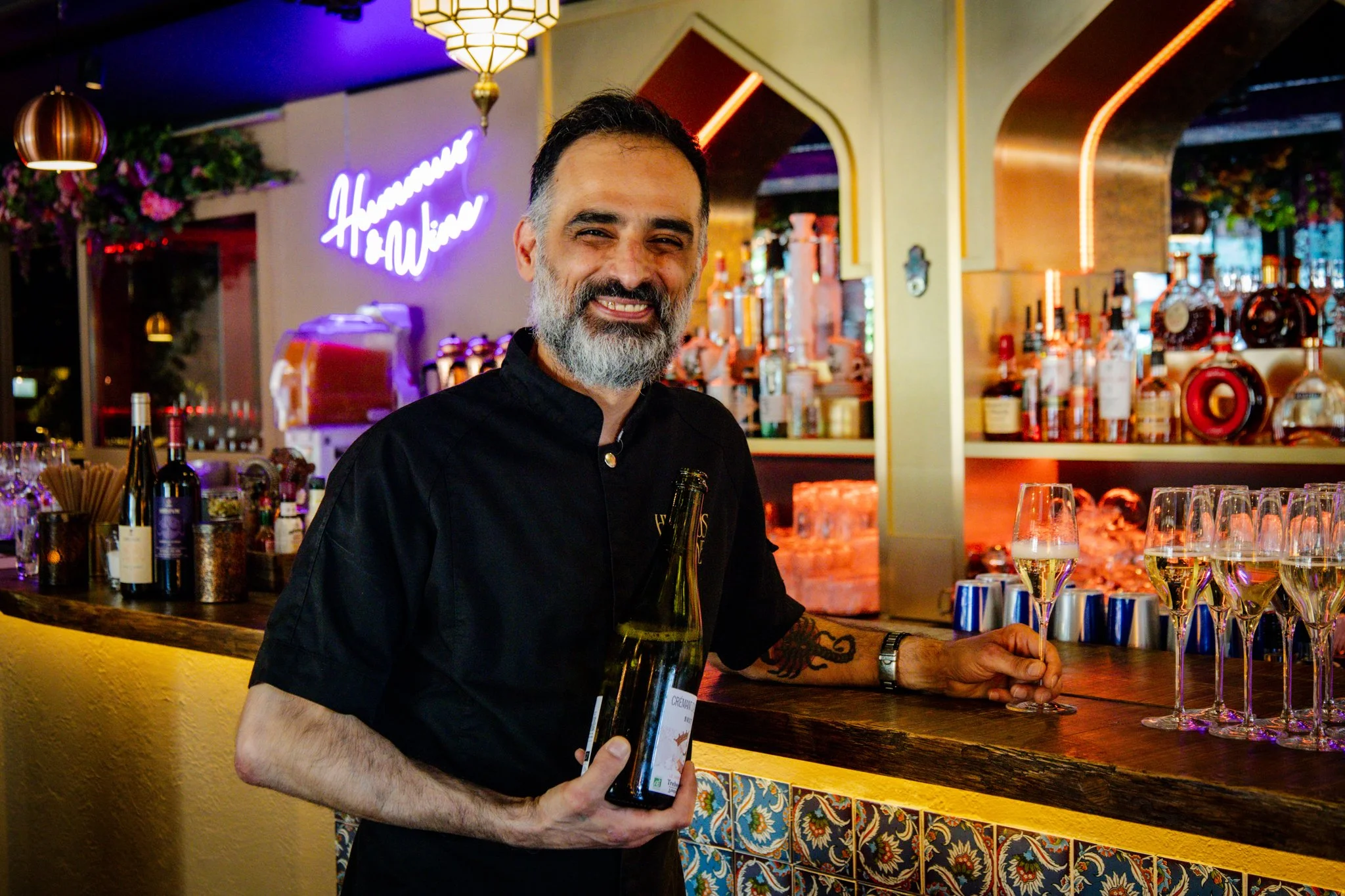 A man with a beard and short dark hair, wearing a black shirt, smiling and holding a bottle of wine, standing at a bar with glasses of champagne. The background features colorful lights, bottles, and a neon sign in a lively bar or restaurant setting.